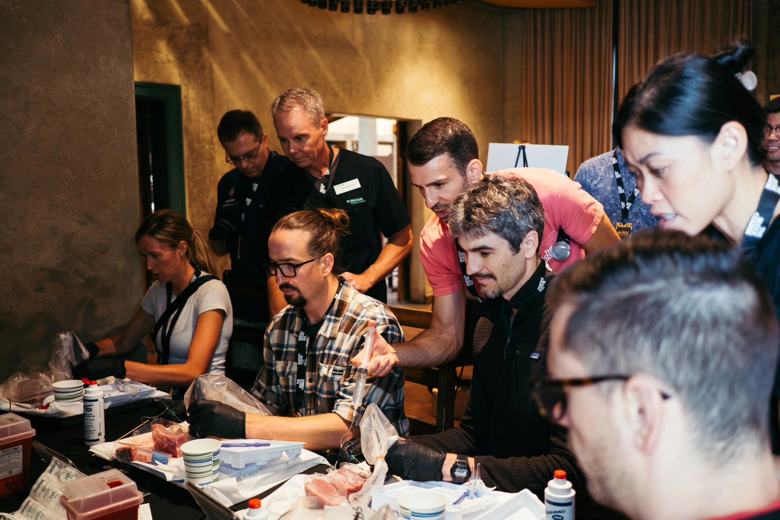 Group of people gathered around a table during a medical training or workshop, with some participants wearing gloves and handling medical supplies.