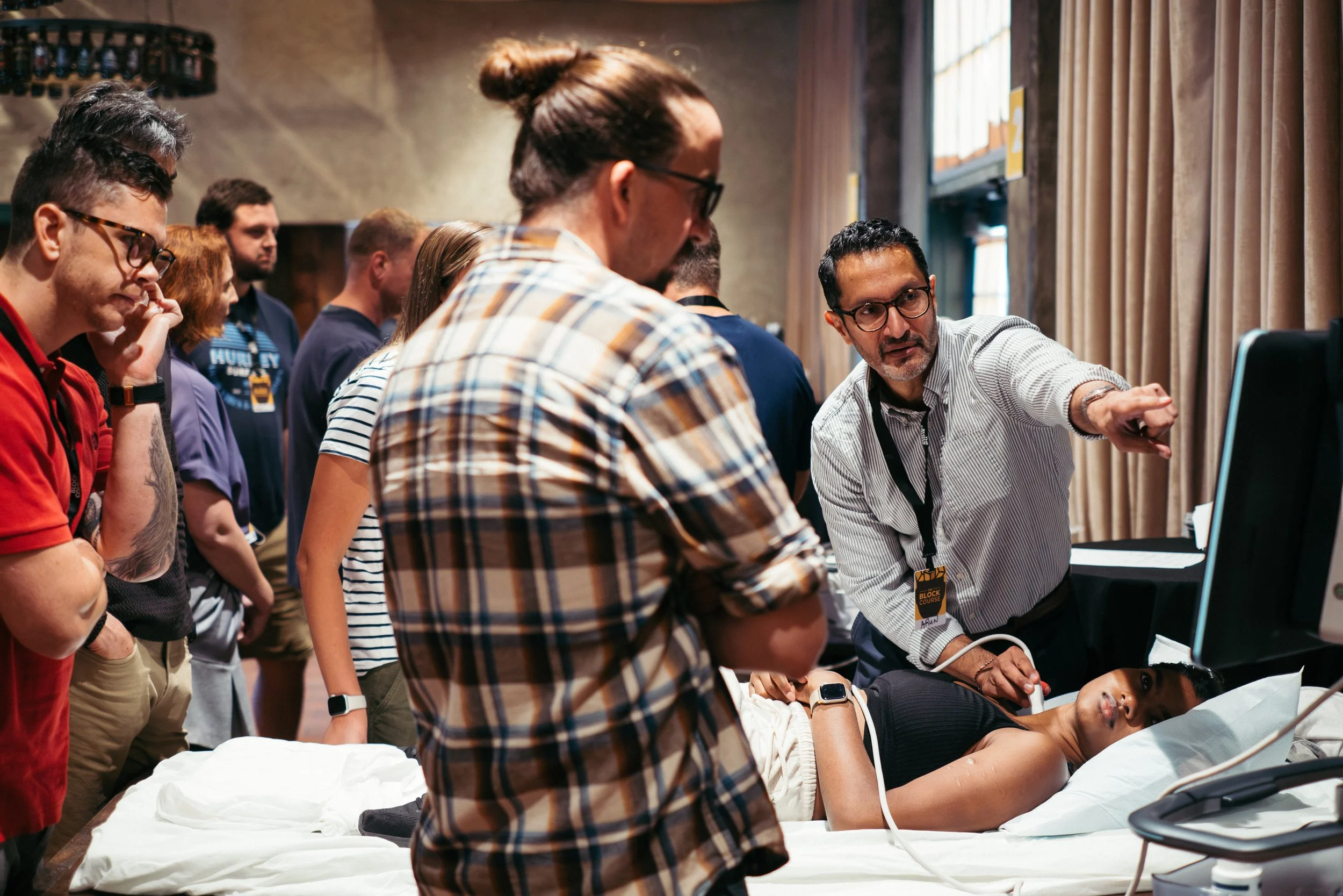 Medical demonstration or training with a group of people gathered around a patient lying on a hospital bed, with a presenter showing medical equipment and explaining.