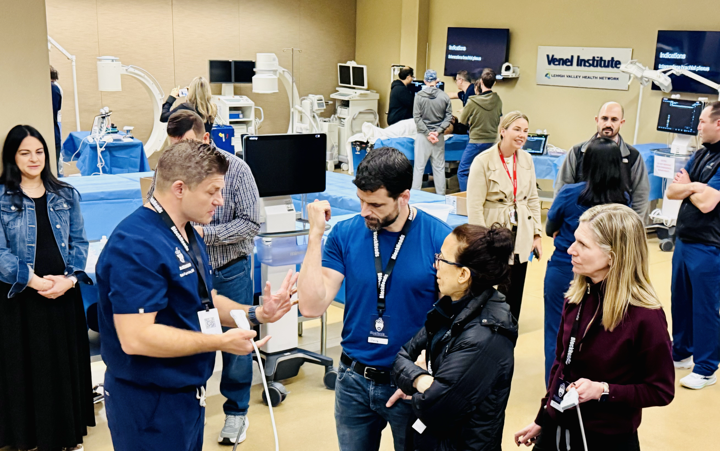 Medical professionals and staff gathered in a hospital or medical training room with medical equipment, monitors, and beds, engaging in discussion and observing medical procedures.