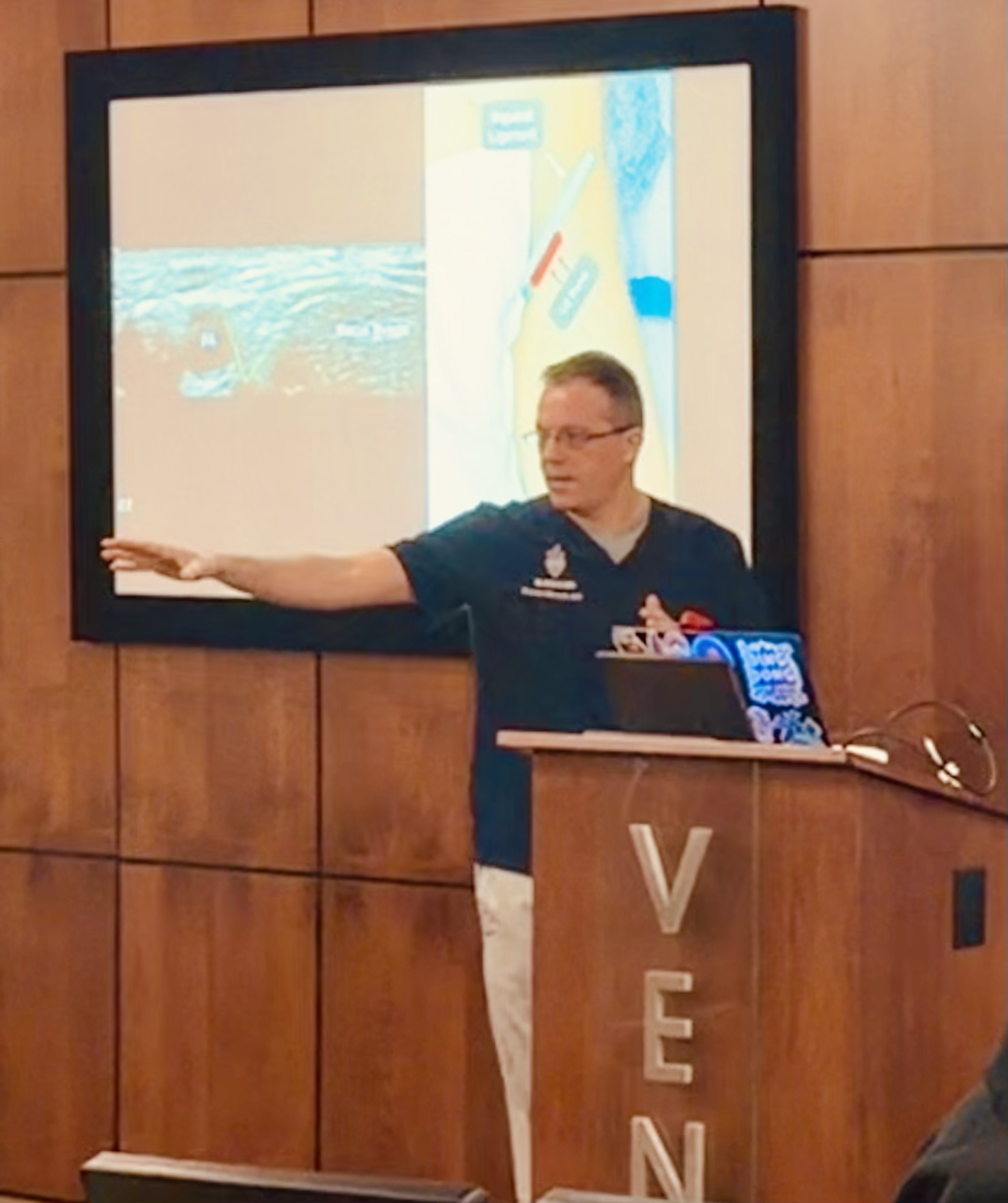 A man in a dark uniform giving a presentation in front of a large screen displaying a diagram of an aircraft's interior, with wooden panel walls in the background.