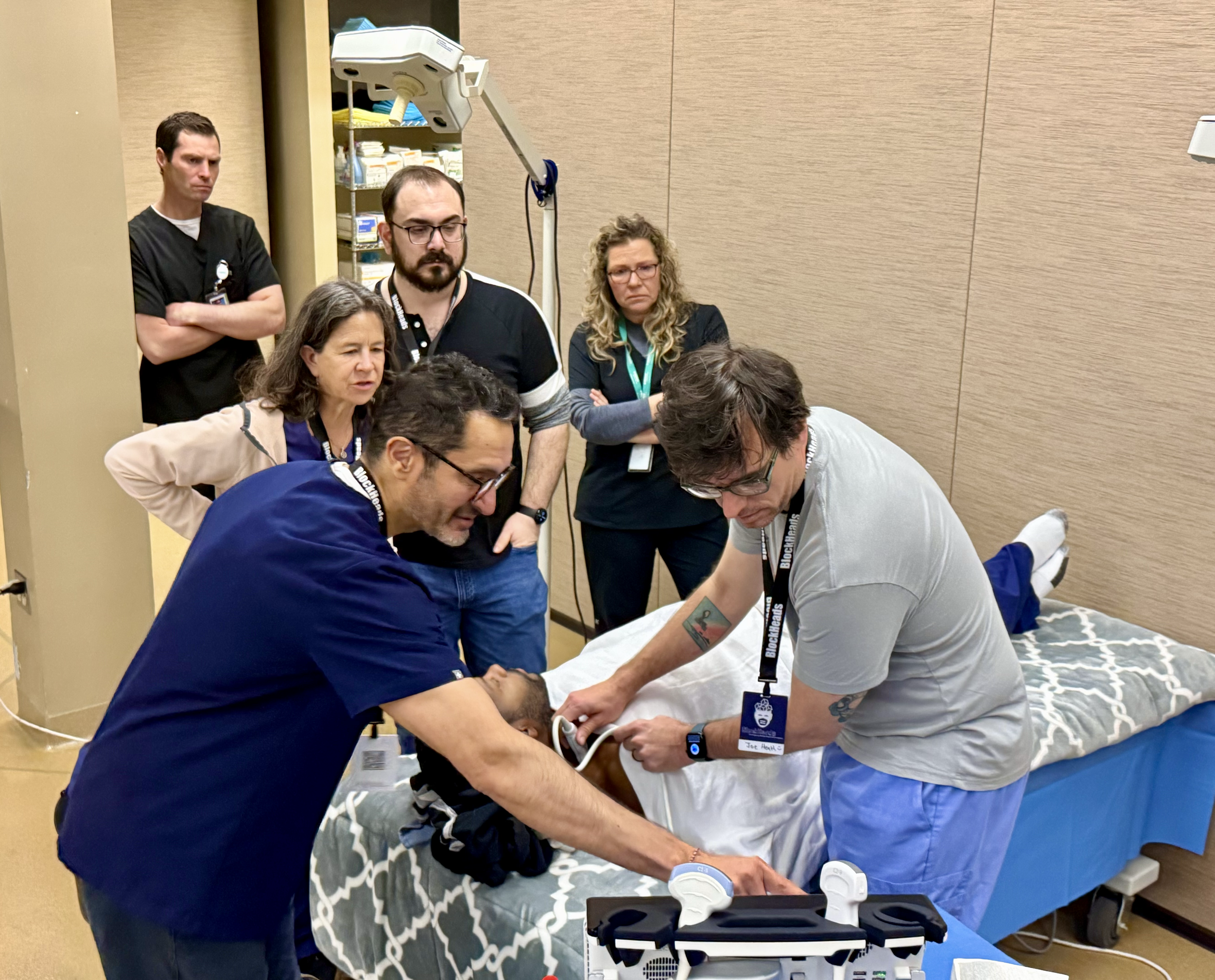 Medical professionals attending to a patient lying on a hospital bed, with several people observing. One person is using medical equipment on the patient, while others watch attentively.