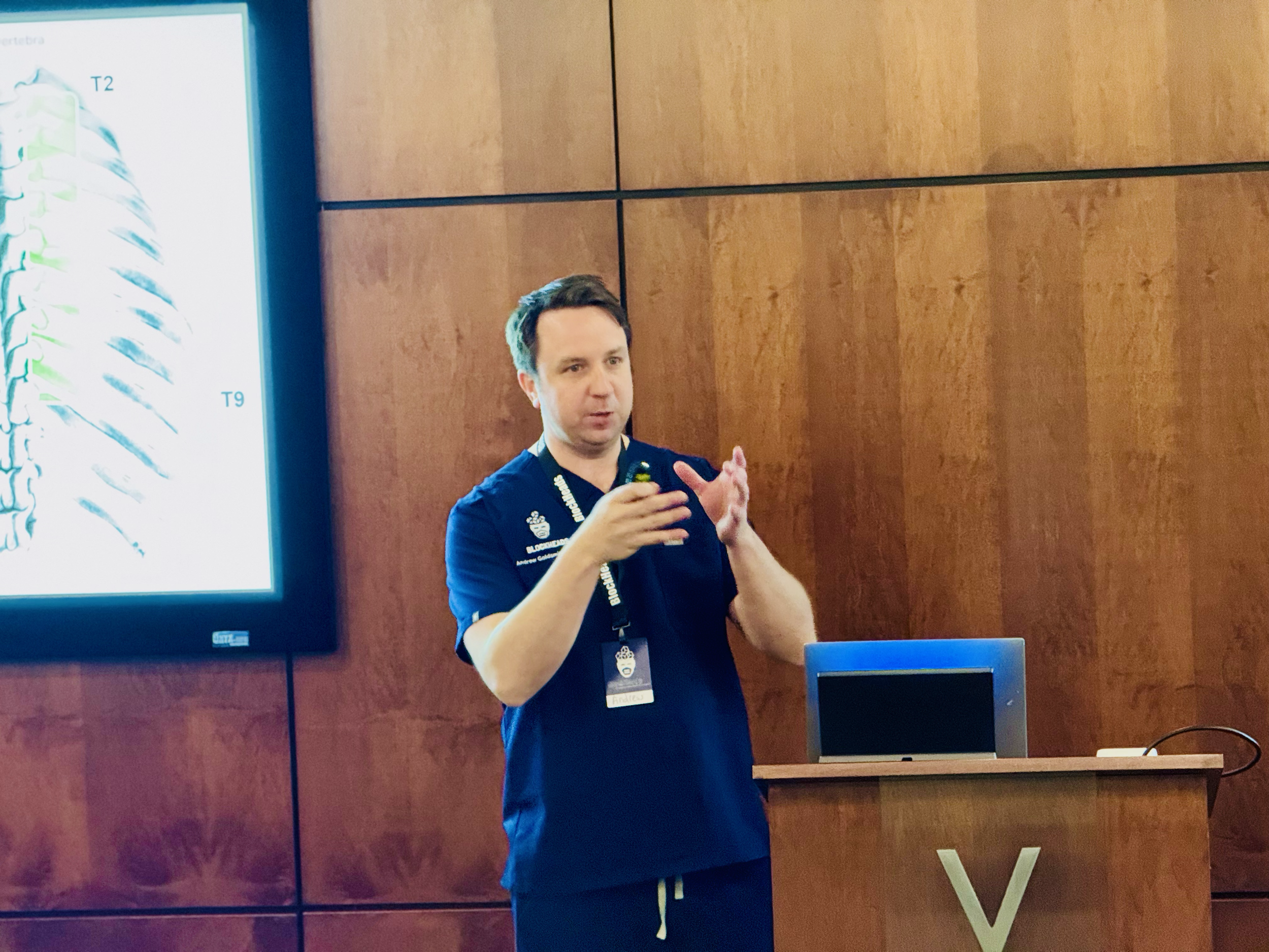 Man giving a presentation in front of a wooden wall, with a large screen displaying a diagram of a spinal column.