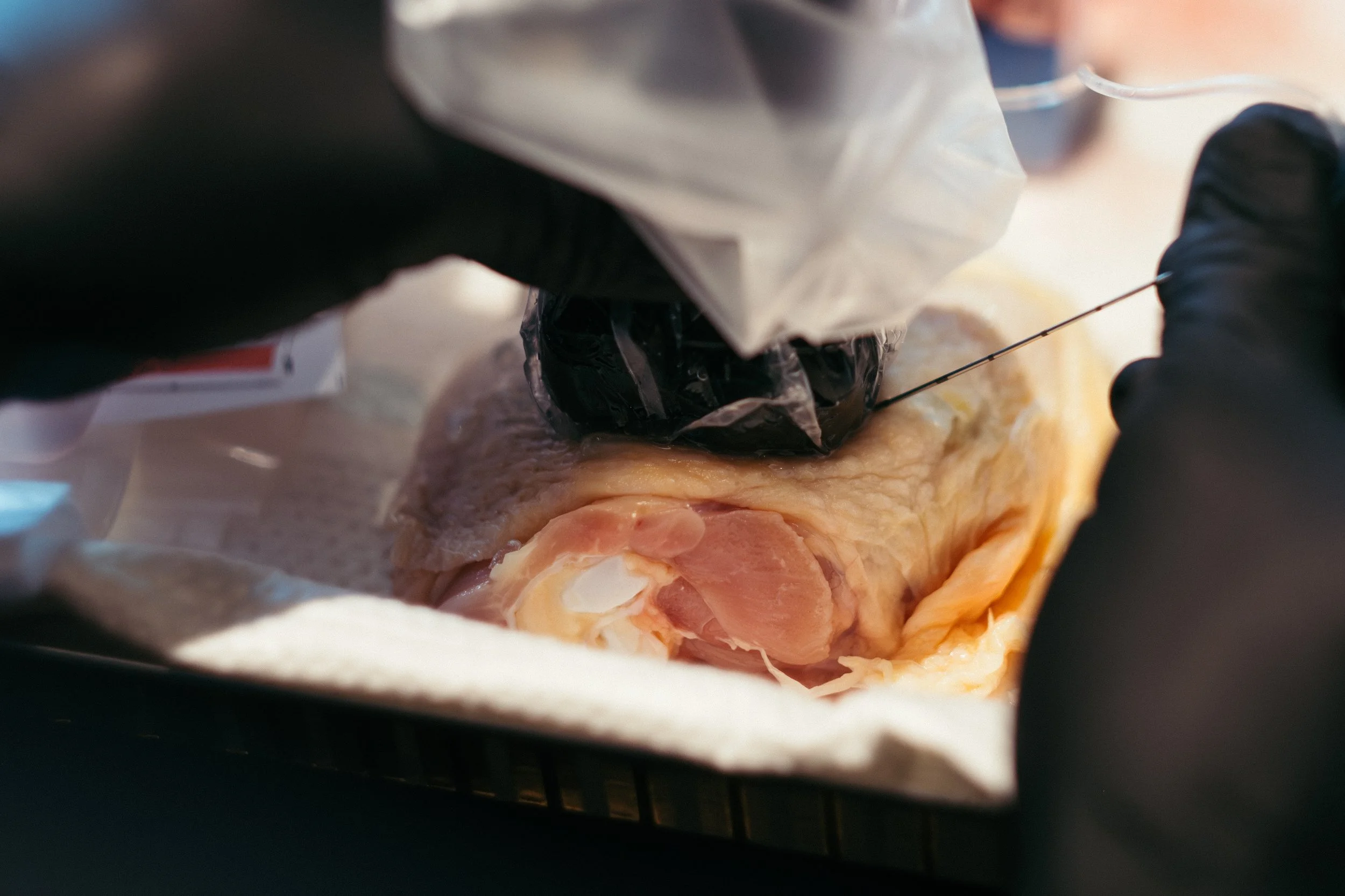 Close-up of a person in black gloves injecting substances into a raw chicken with a syringe and vacuum-sealed bag.