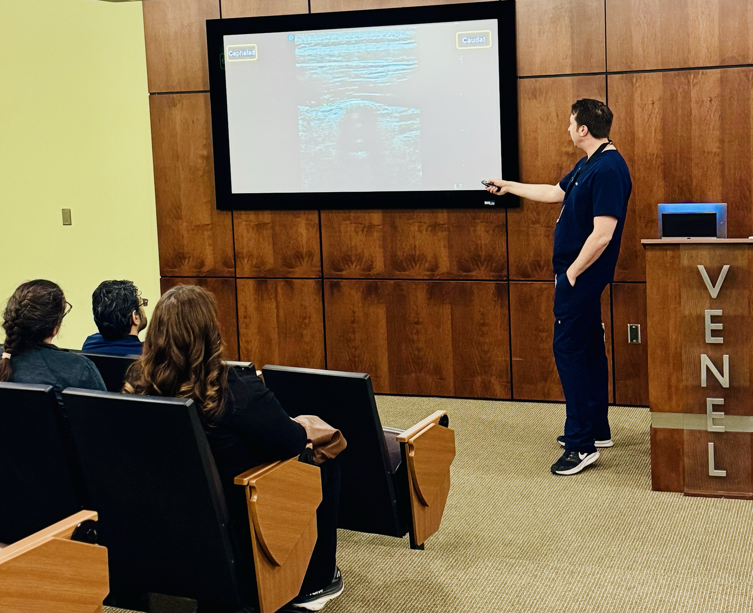 Medical professional giving a presentation on an ultrasound image in a conference room with seated audience.