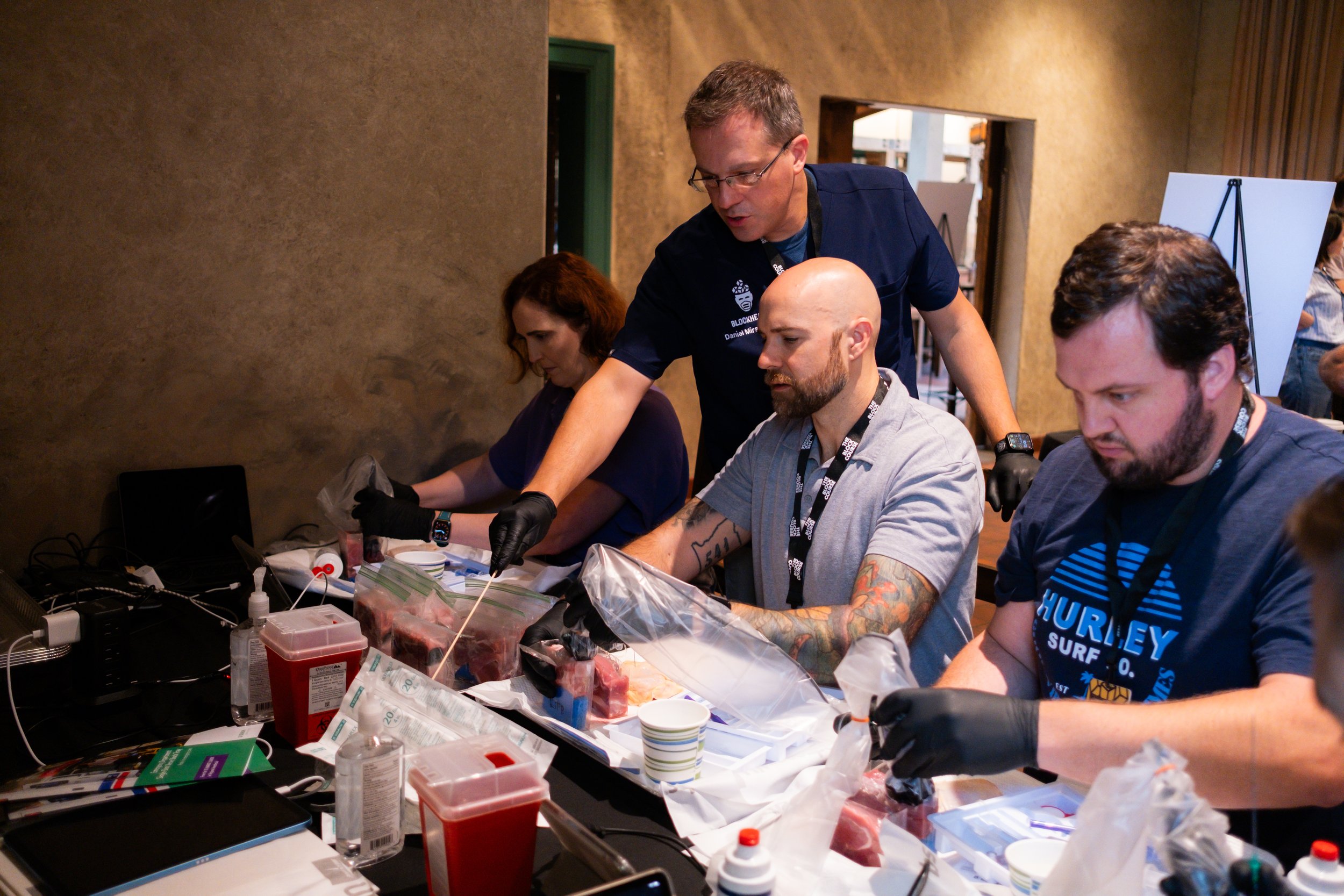 A group of people in a workshop handling raw meat, with one person in a blue shirt explaining or demonstrating to others.