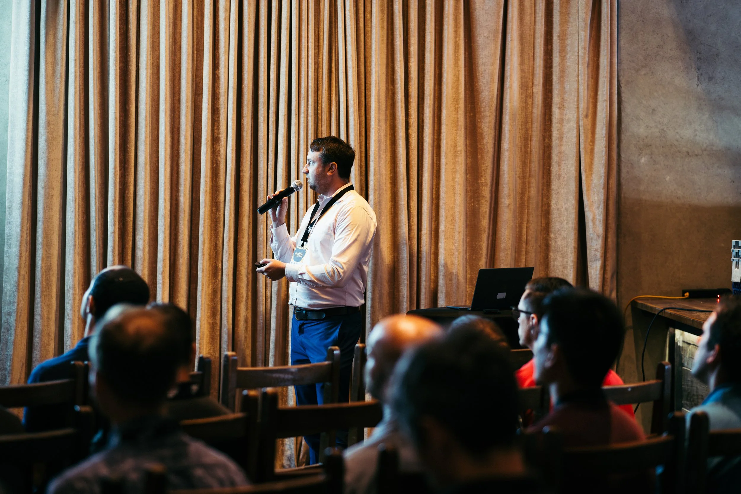 A man giving a presentation with a microphone to an audience in a conference room with gold curtains.