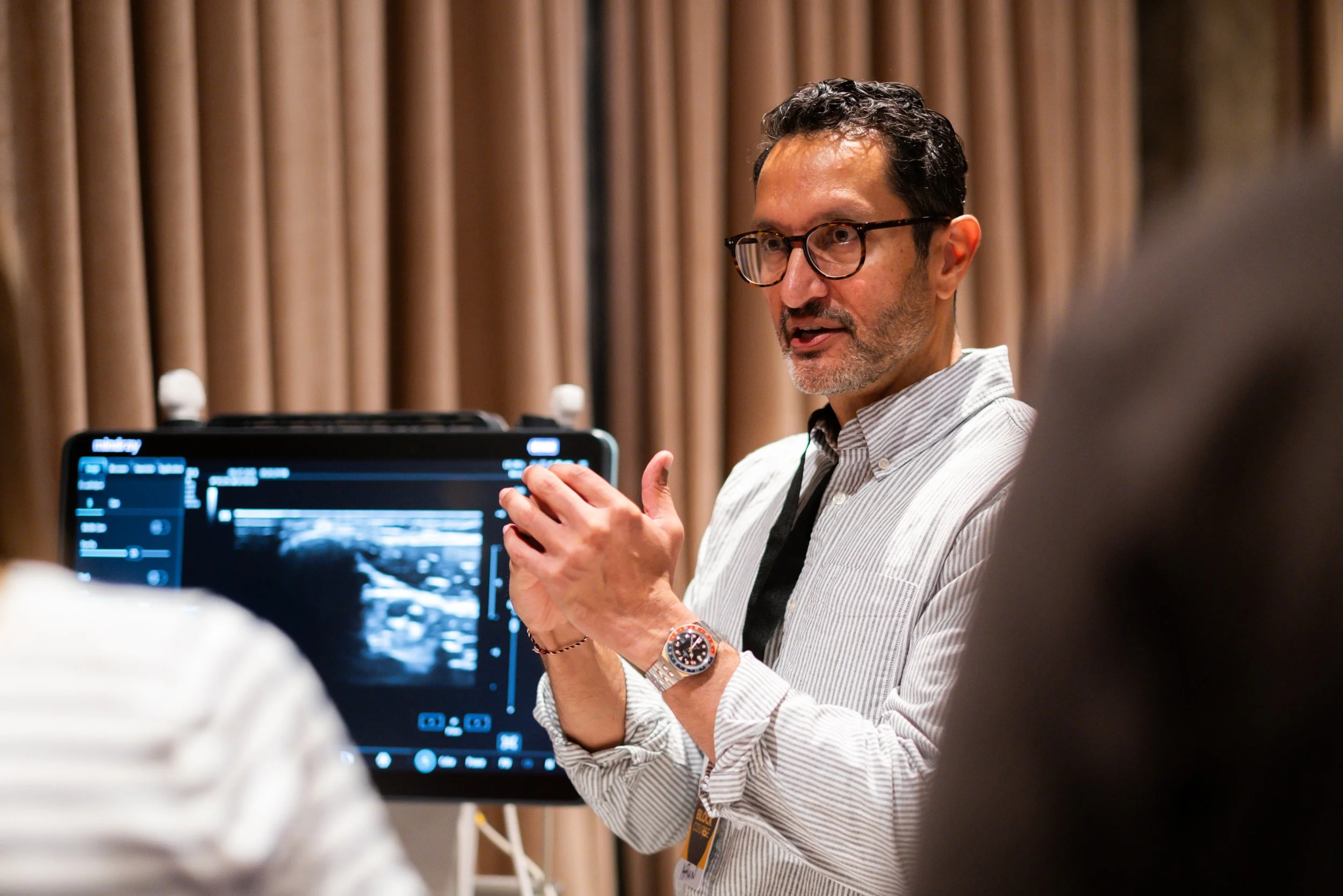 A man with glasses and a watch explains ultrasound images to a group in a conference room with brown curtains.