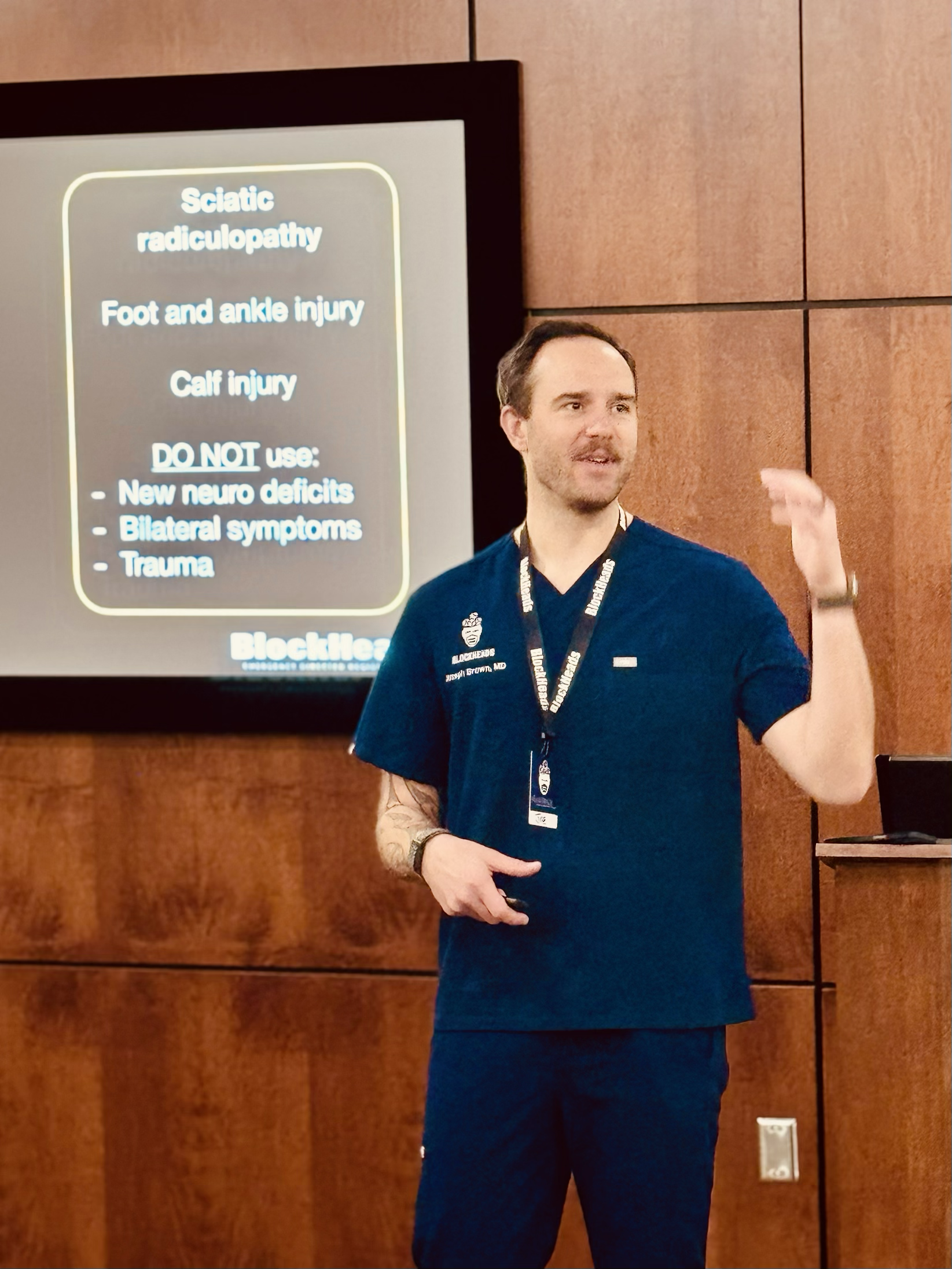 Medical professional giving a presentation on sciatic radiculopathy, foot and ankle injury, and calf injury, standing in front of a screen with related information, inside a room with wooden walls.