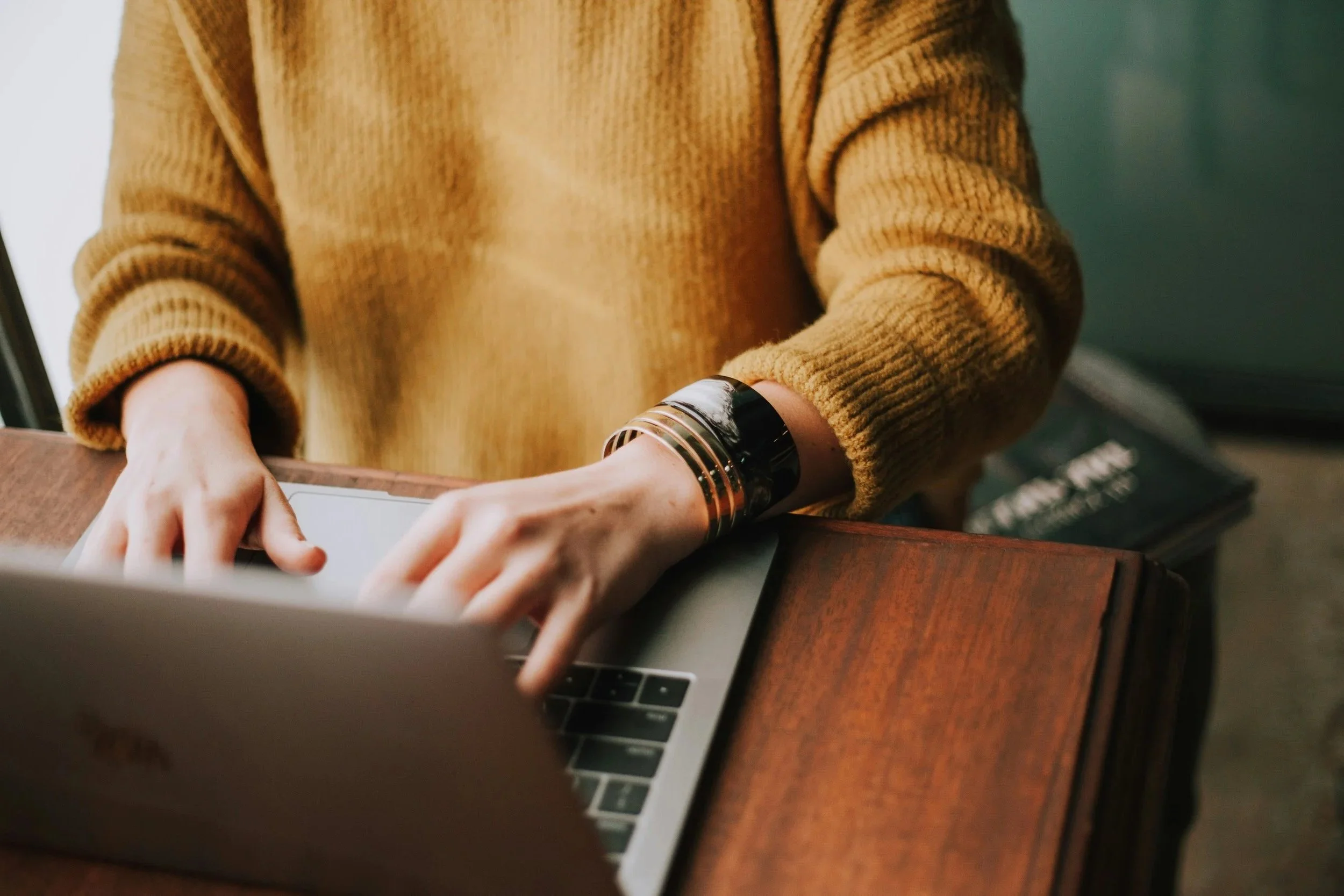 Person wearing a mustard-colored sweater using a laptop on a wooden table, with a stack of magazines or books nearby.