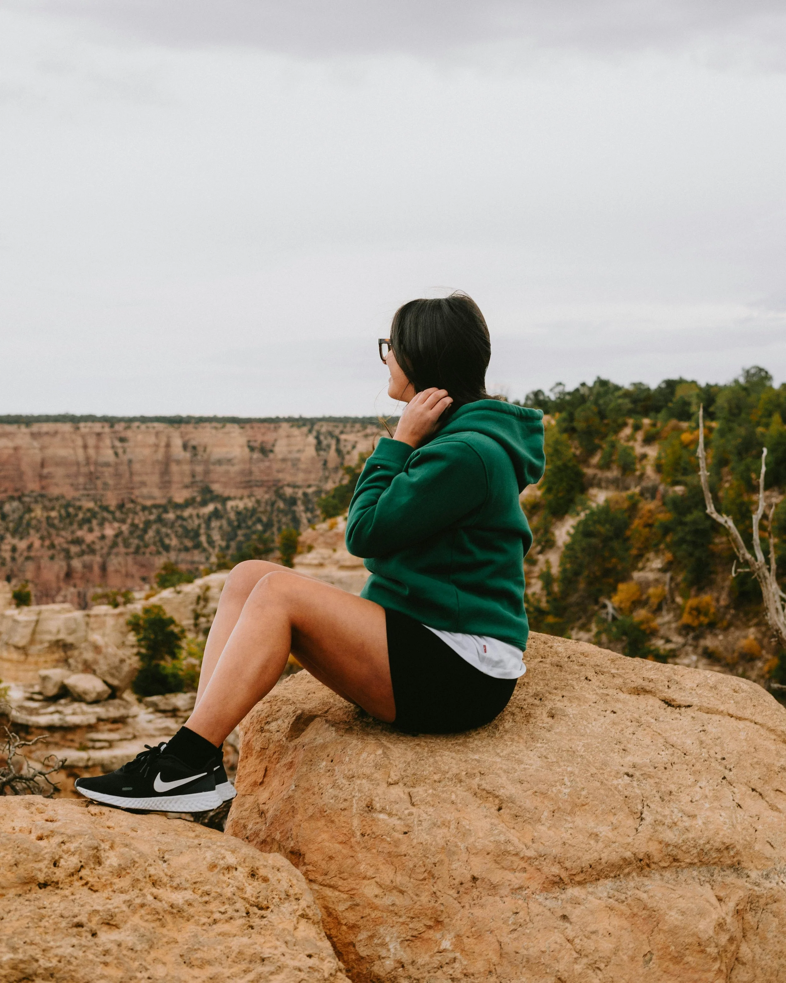 Woman sitting on a large rock in a canyon, facing away, wearing a green hoodie, black shorts, and sneakers, with trees and cliffs in the background.
