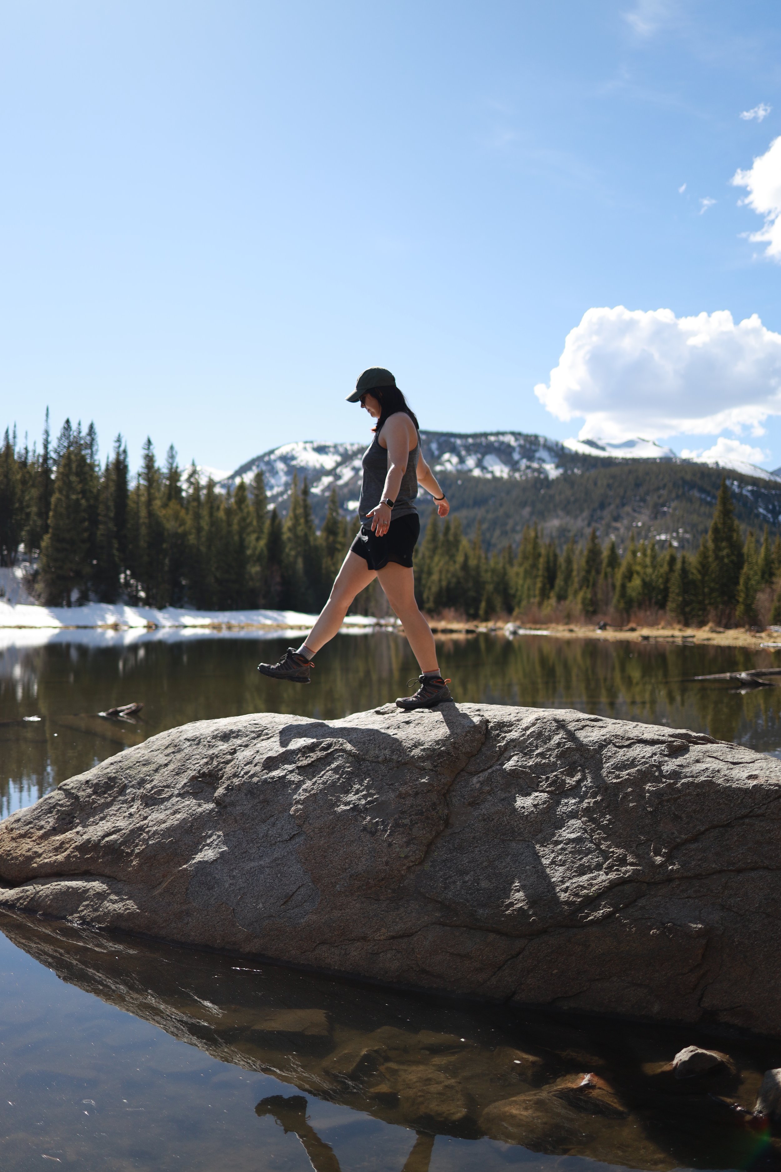 A woman wearing a hat, sunglasses, gray tank top, and black shorts balancing while walking on a large rock over a calm lake in a mountainous landscape with snow patches, green trees, and blue sky with some clouds.