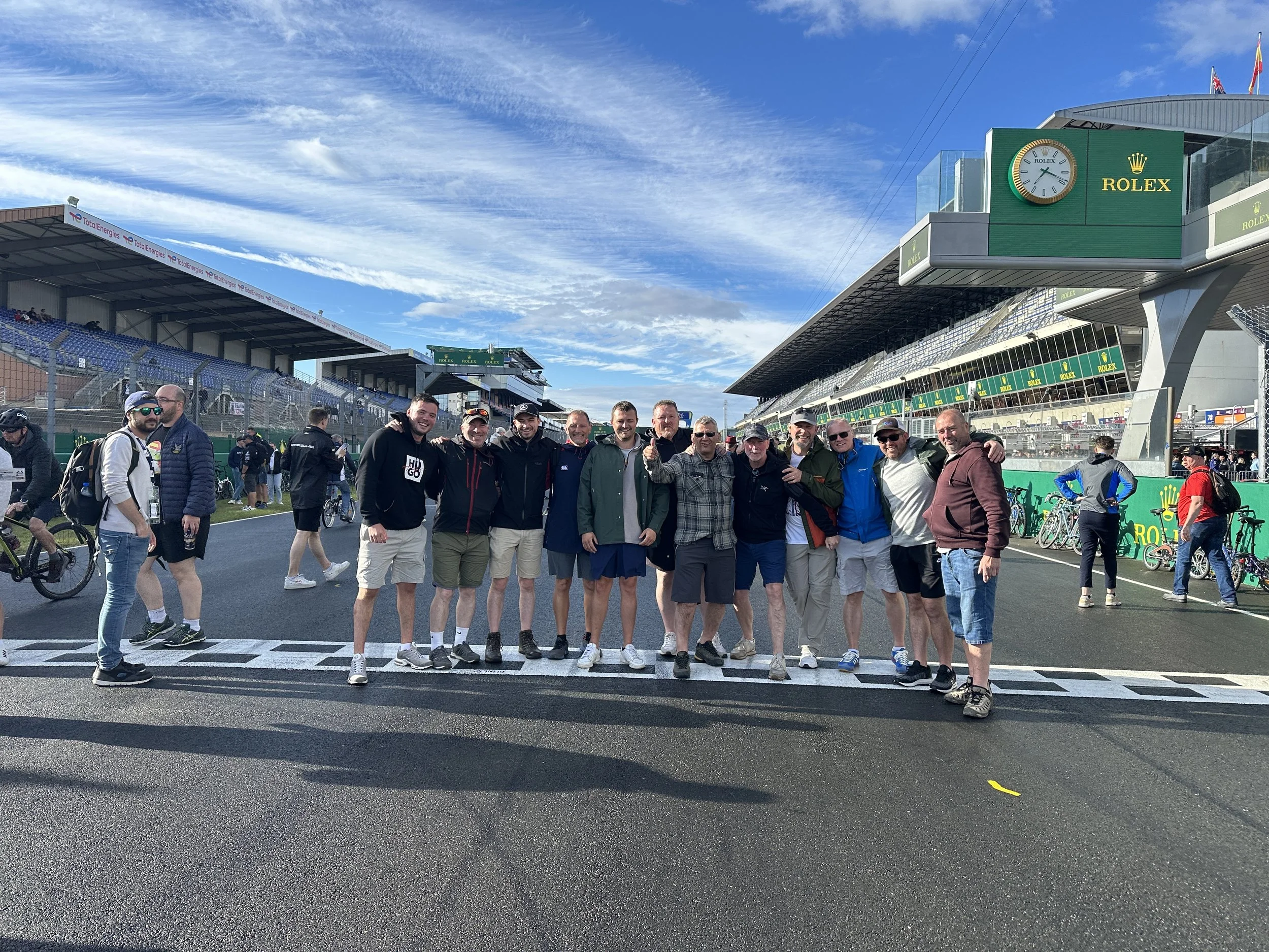 A group of people standing on a race track at a motorsport event, with spectators in the background and a prominent clock displaying the Rolex logo. The sky is partly cloudy.