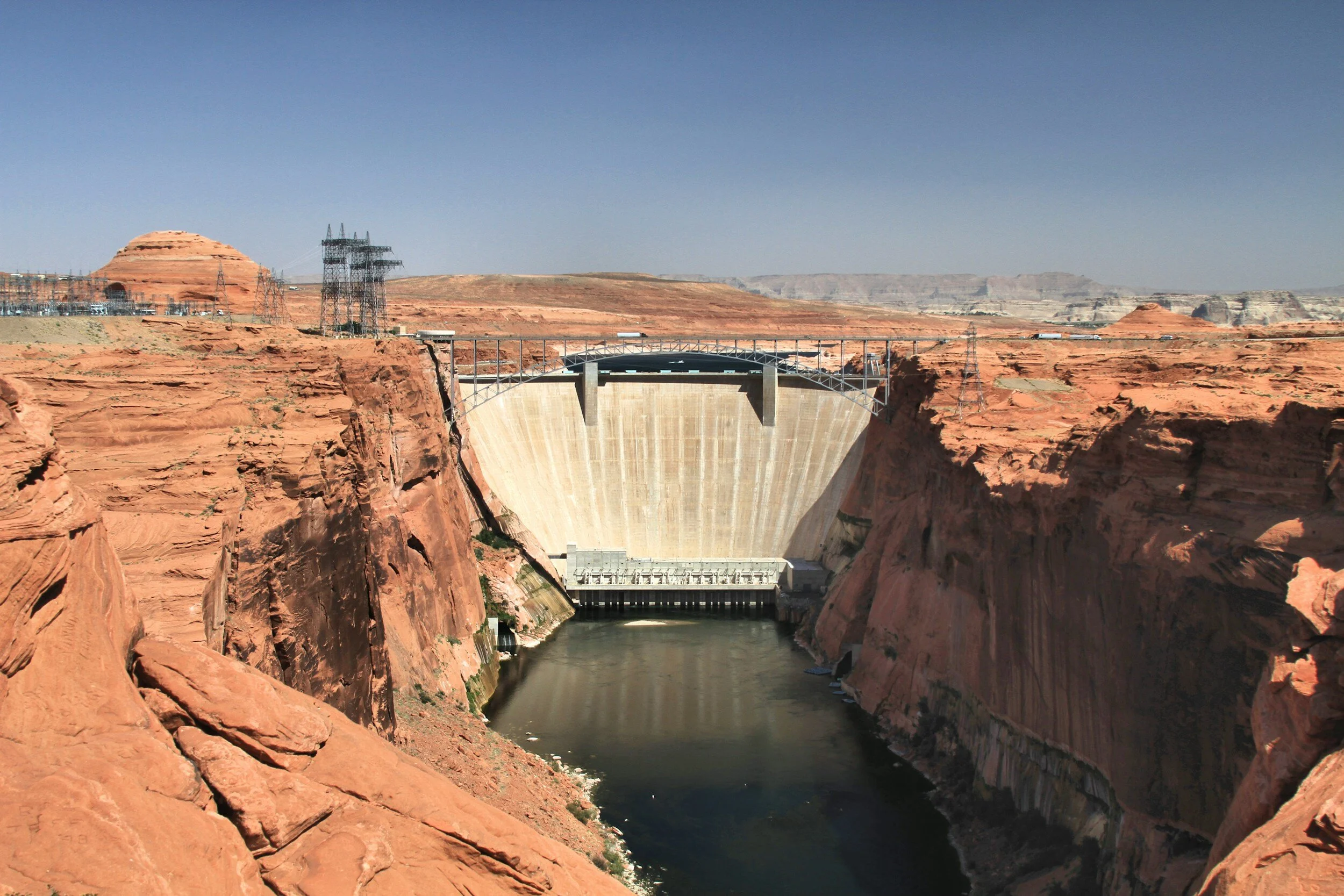 Large concrete dam situated between red rock cliffs, with a river flowing below and a blue sky above; electrical infrastructure visible on the left.