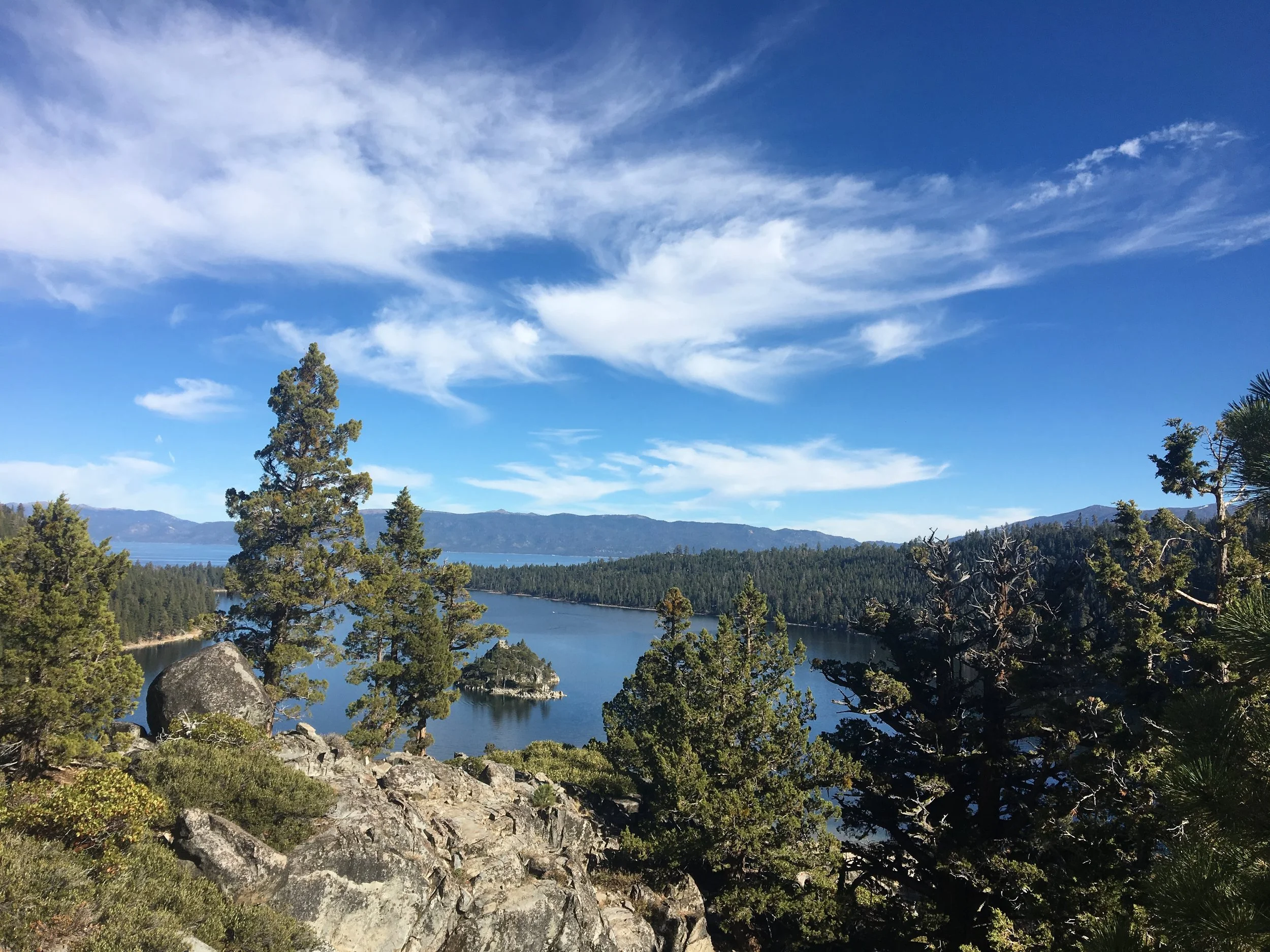 Scenic view of a lake surrounded by evergreen trees, rocky terrain, and distant mountains under a blue sky with wispy clouds.