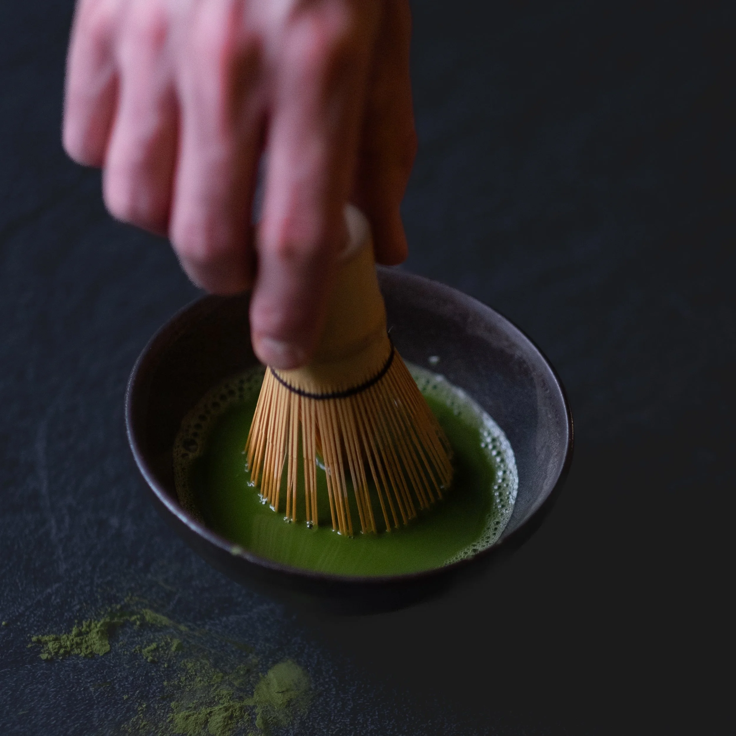 A person's hand is using a bamboo matcha whisk to prepare green tea in a small black bowl on a dark surface.