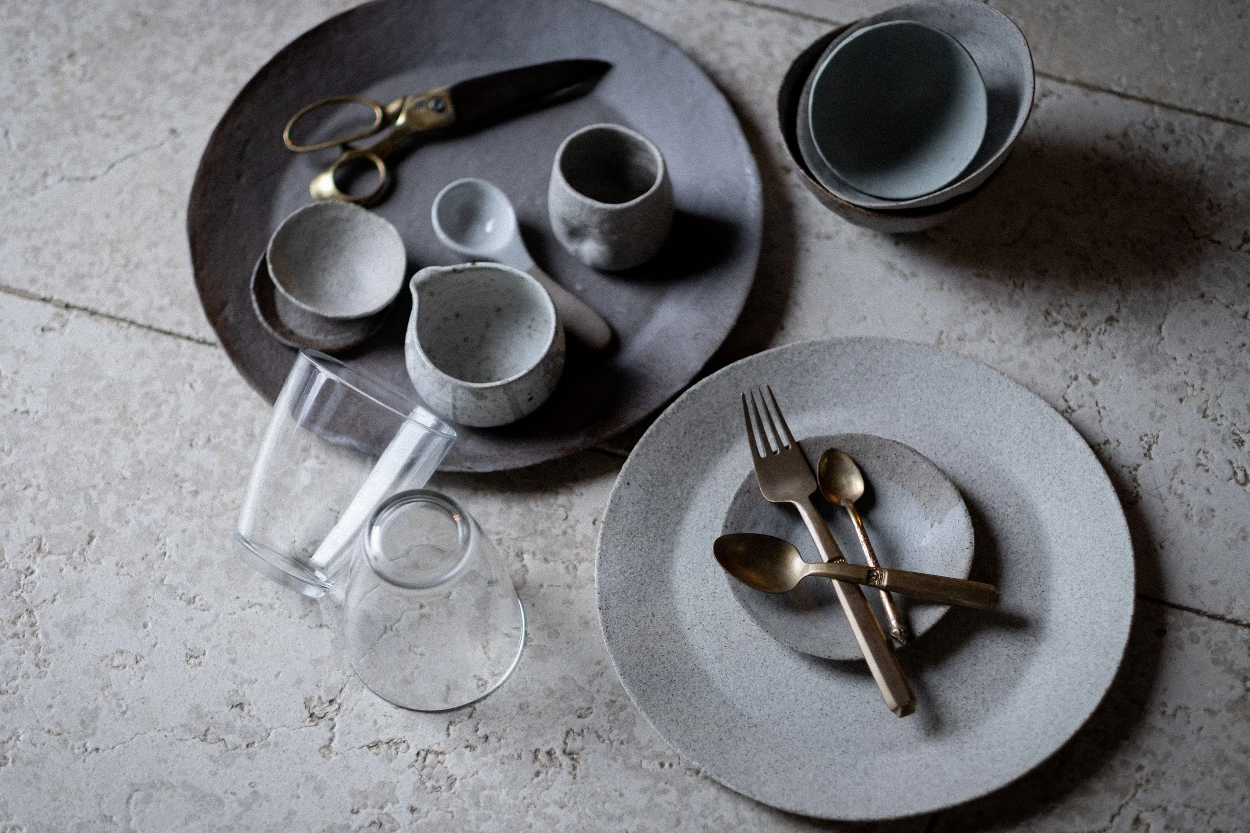 Flat lay of a table setting with ceramic plates, bowls, shot glasses, a fork, a spoon, a pair of scissors, and a small cup, arranged on a textured surface.