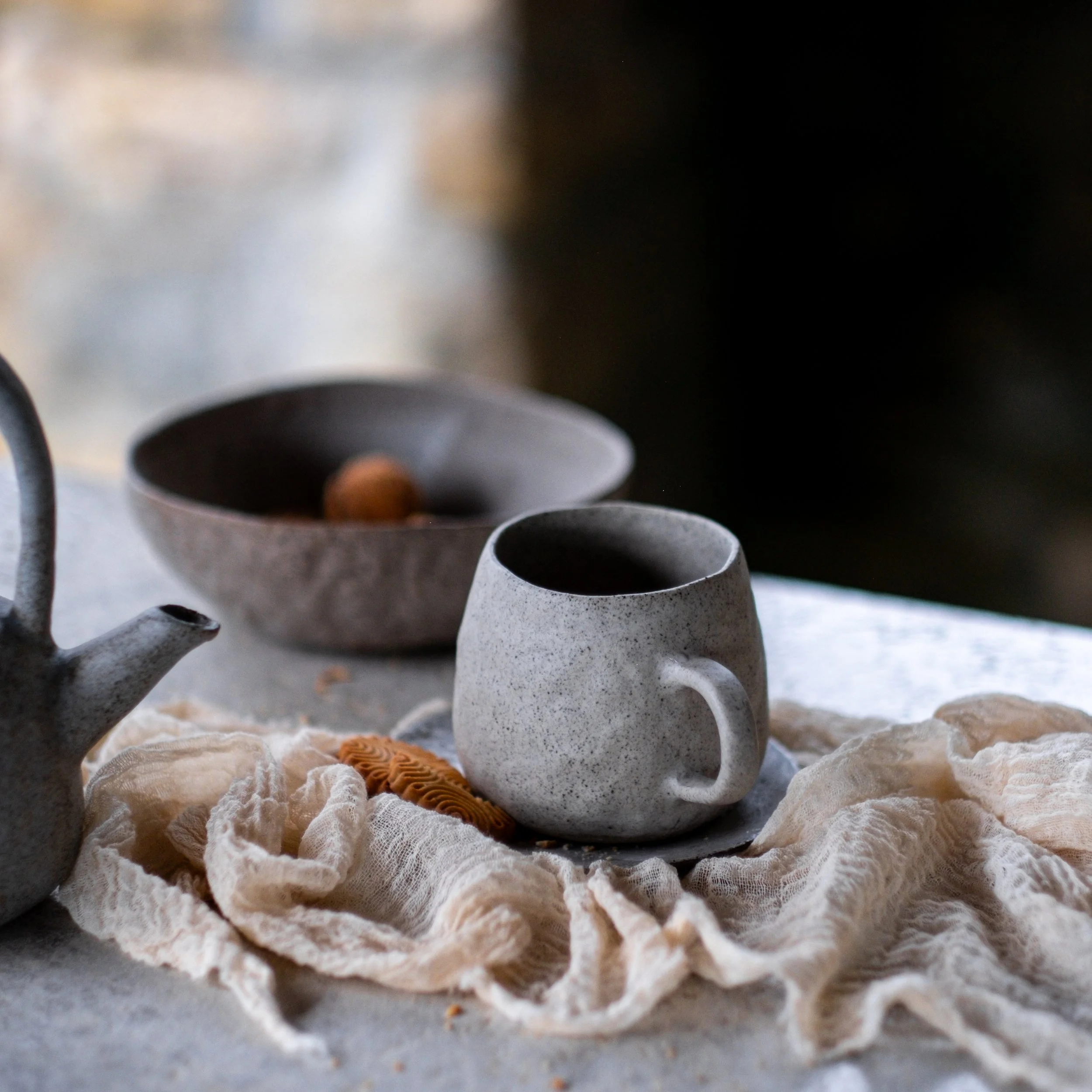 A speckled ceramic mug filled with dark coffee, placed on a light cloth on a table, with a ceramic teapot and a shallow bowl in the background. 