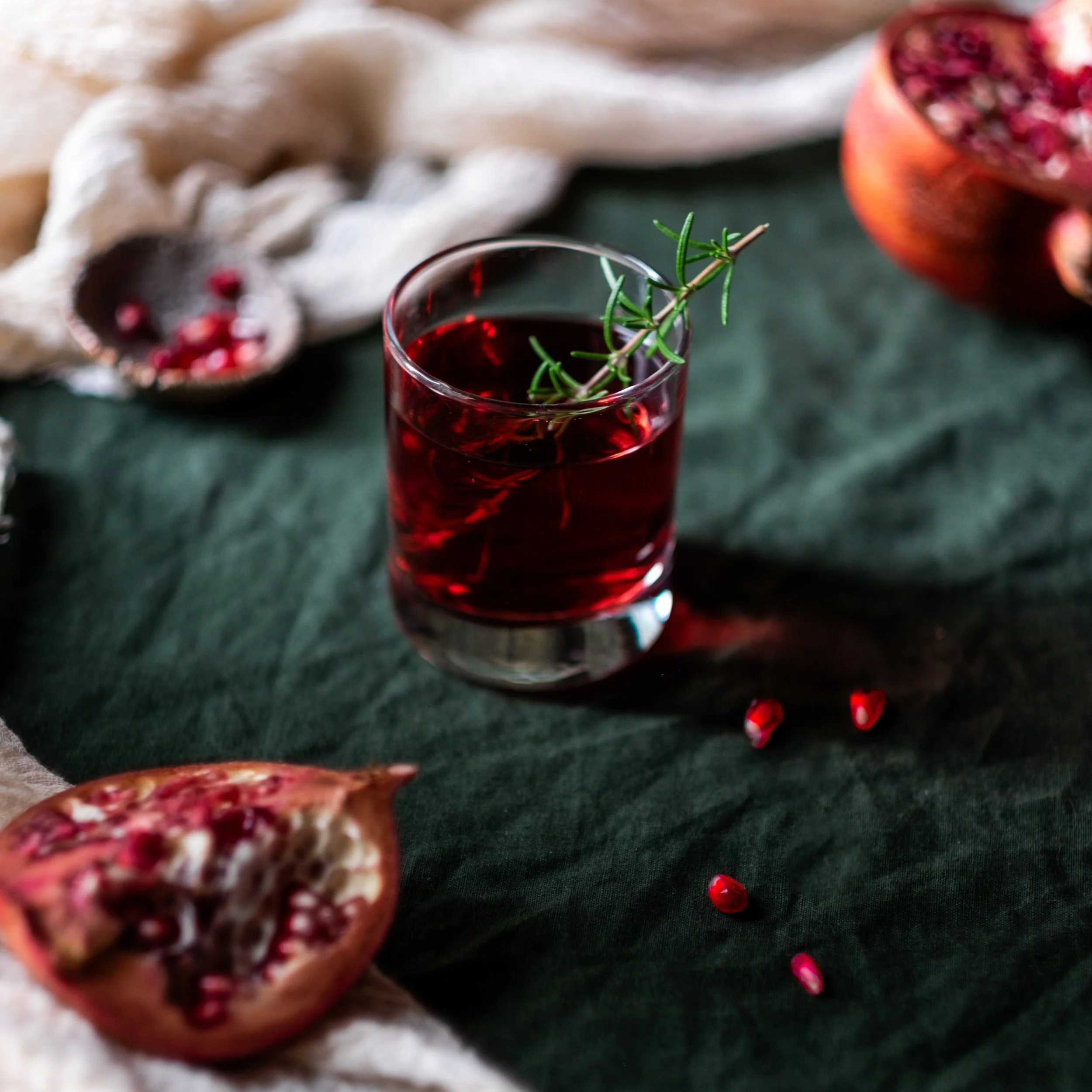 A glass of red pomegranate juice garnished with a sprig of rosemary, placed on a dark green cloth with pomegranate halves and seeds around it.
