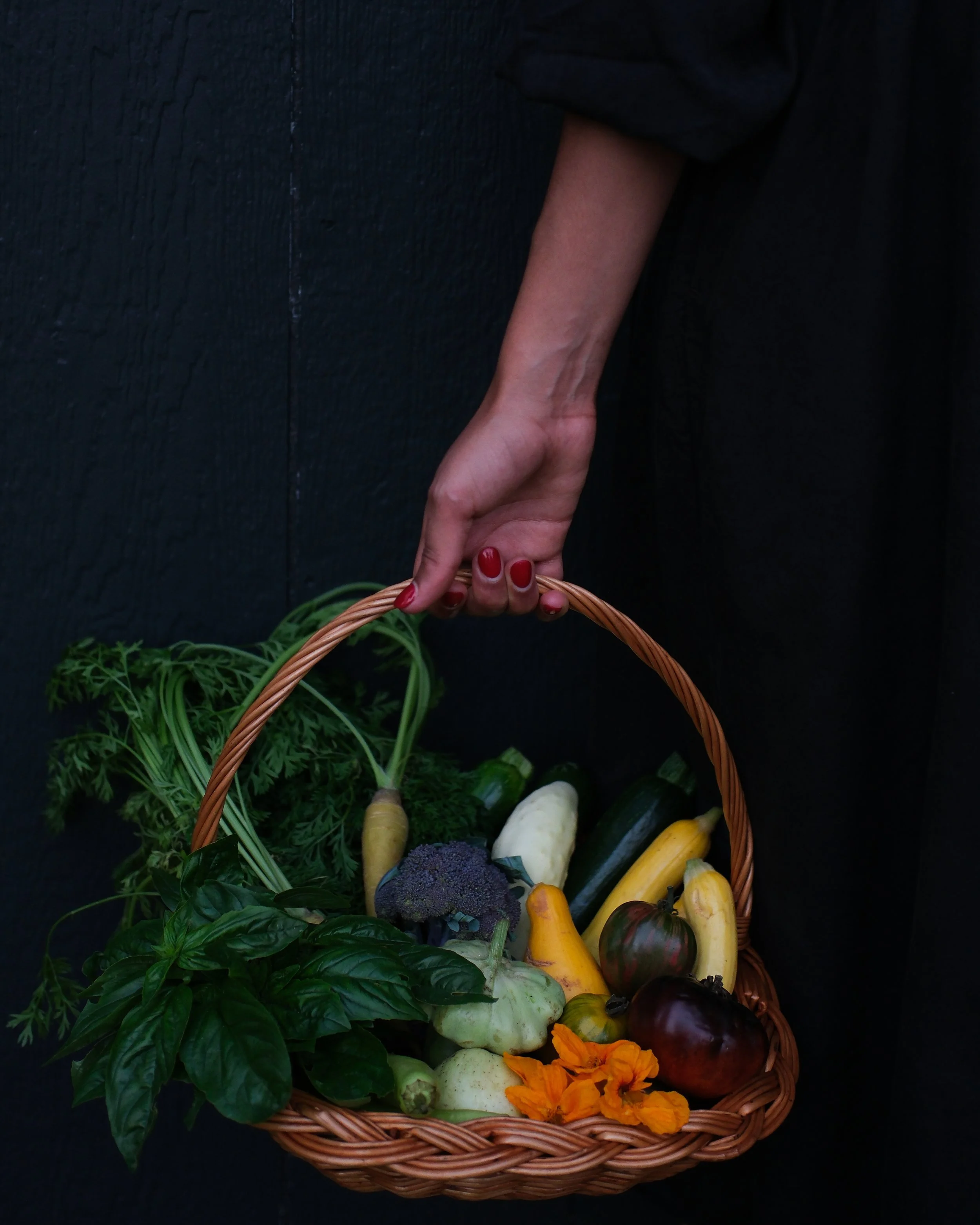 A person's hand with red painted nails holding a basket filled with fresh vegetables and herbs, including zucchini, eggplant, broccoli, parsley, basil, and edible flowers, against a dark background.