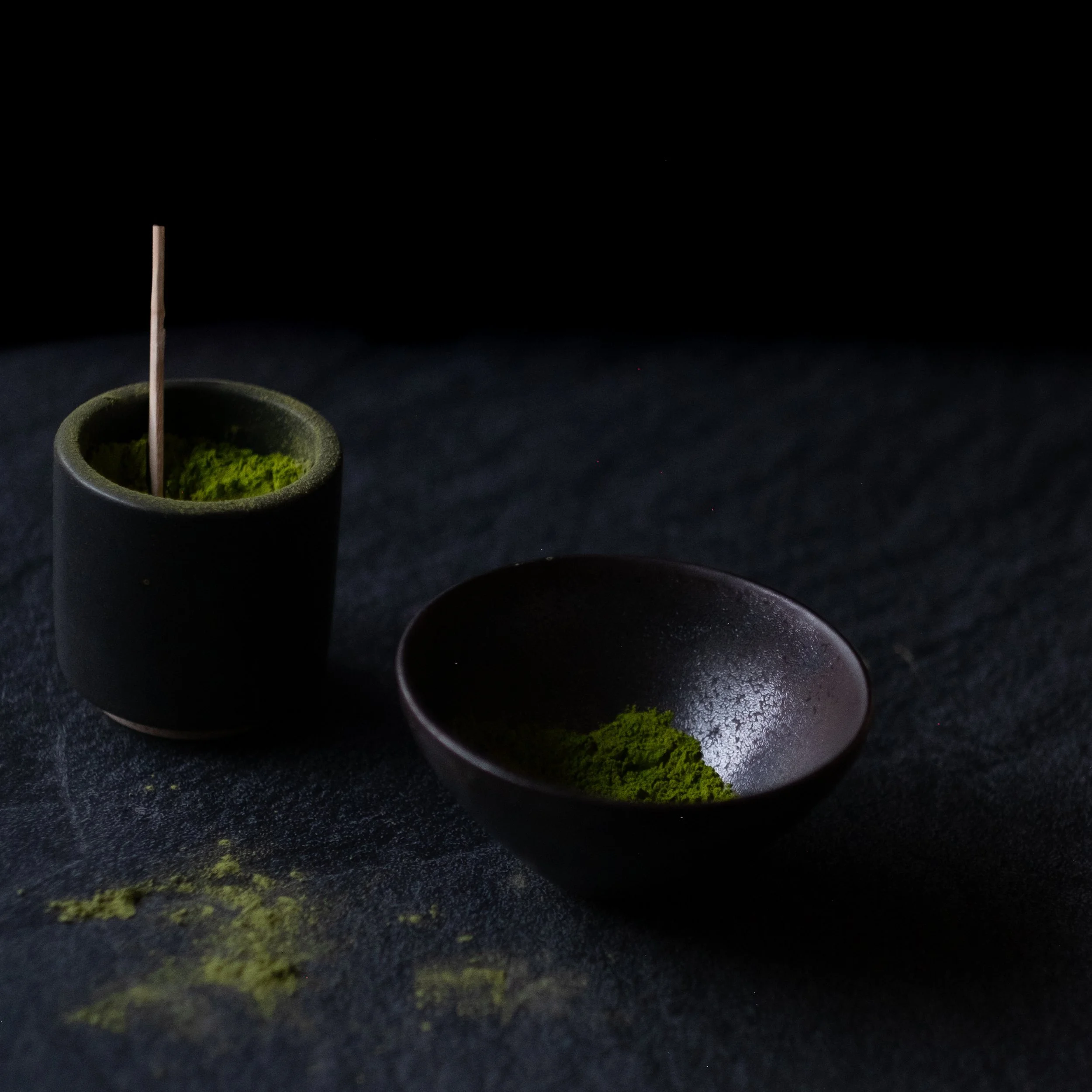 Black bowl and small container filled with green matcha powder on black background.