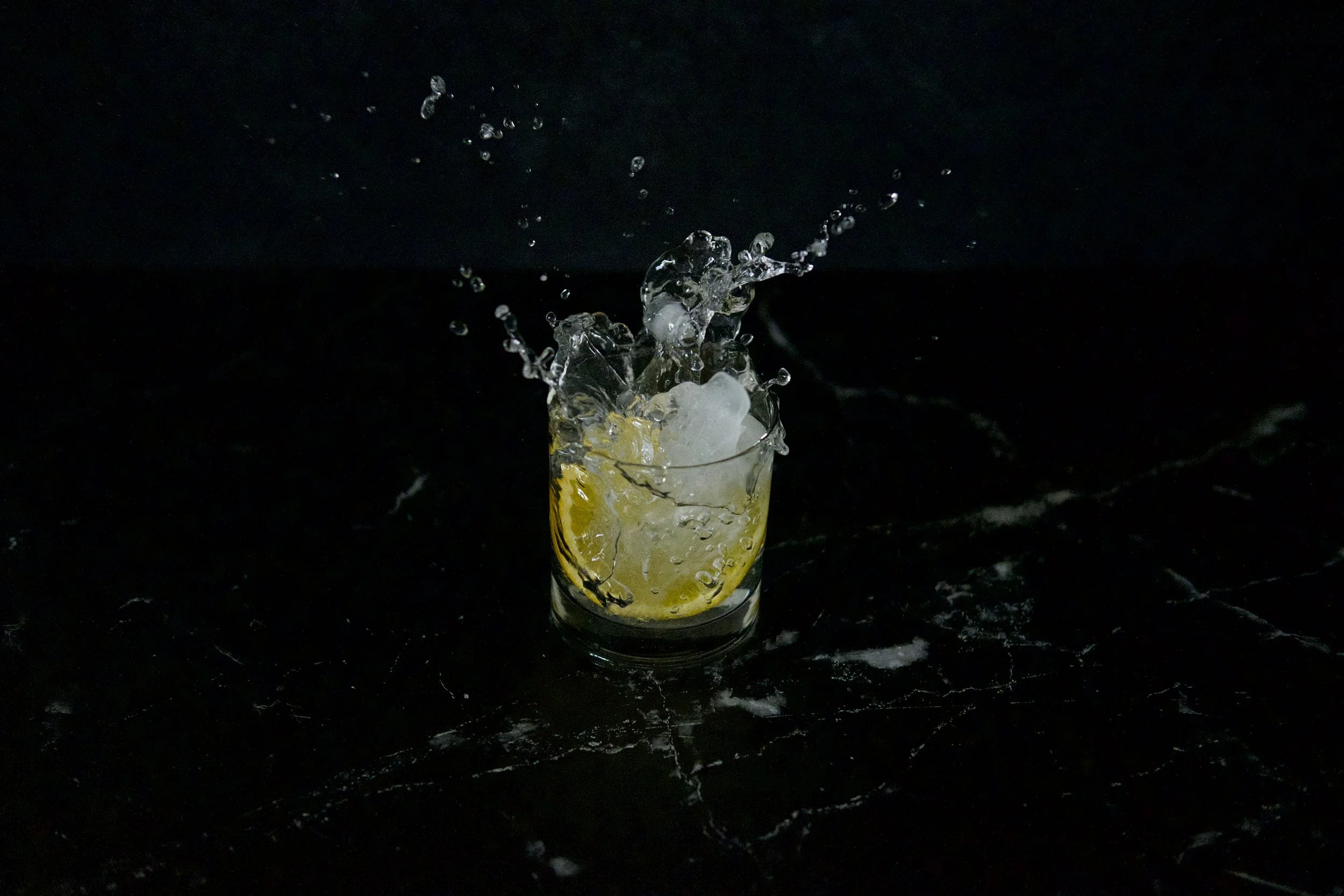 A glass of lemon-infused water with ice, splashing as a lemon slice is dropped into it against a dark background.