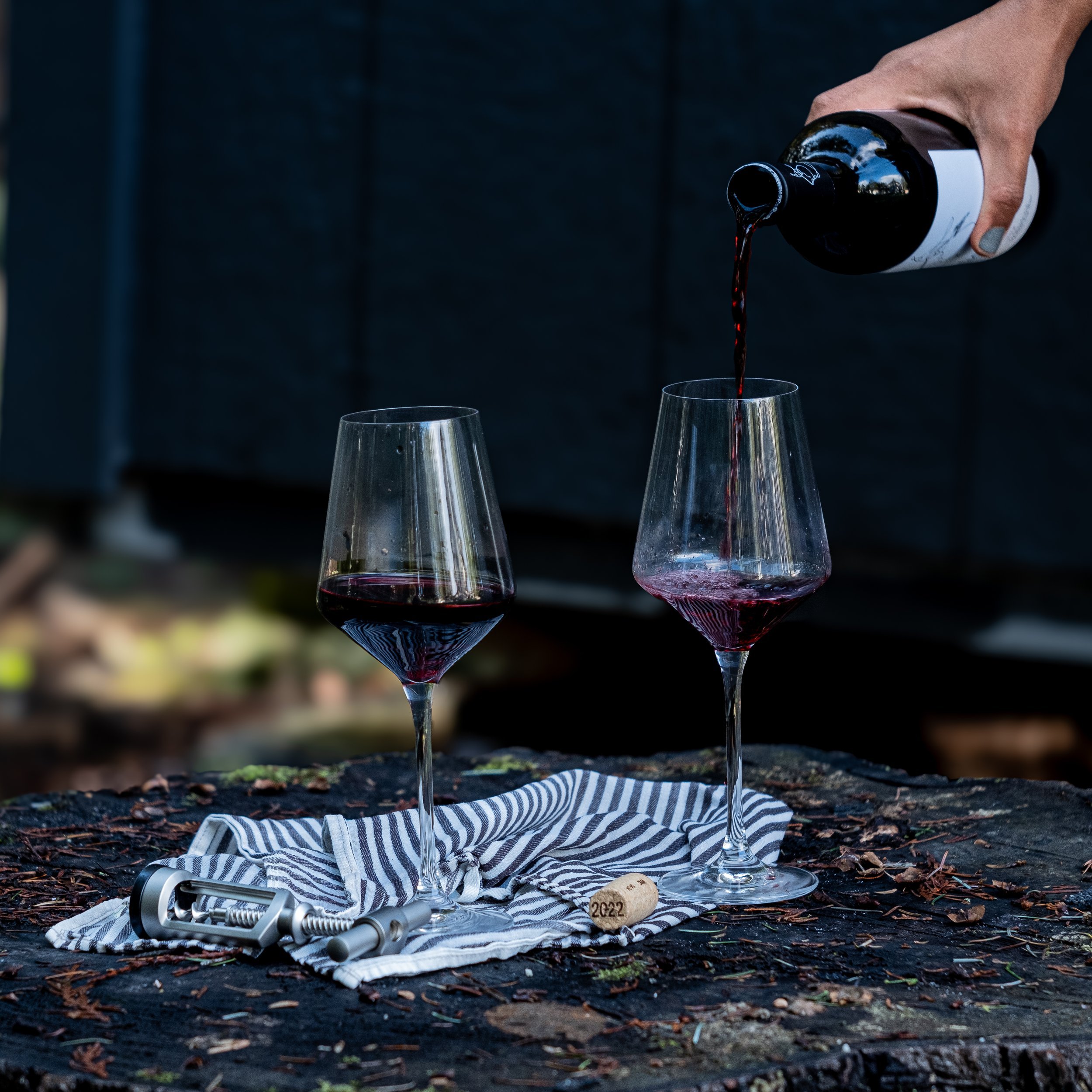 A person pouring red wine into a glass on a rustic wooden surface, with another glass already filled nearby, a cork with the year 2022, and a corkscrew placed on a striped cloth.