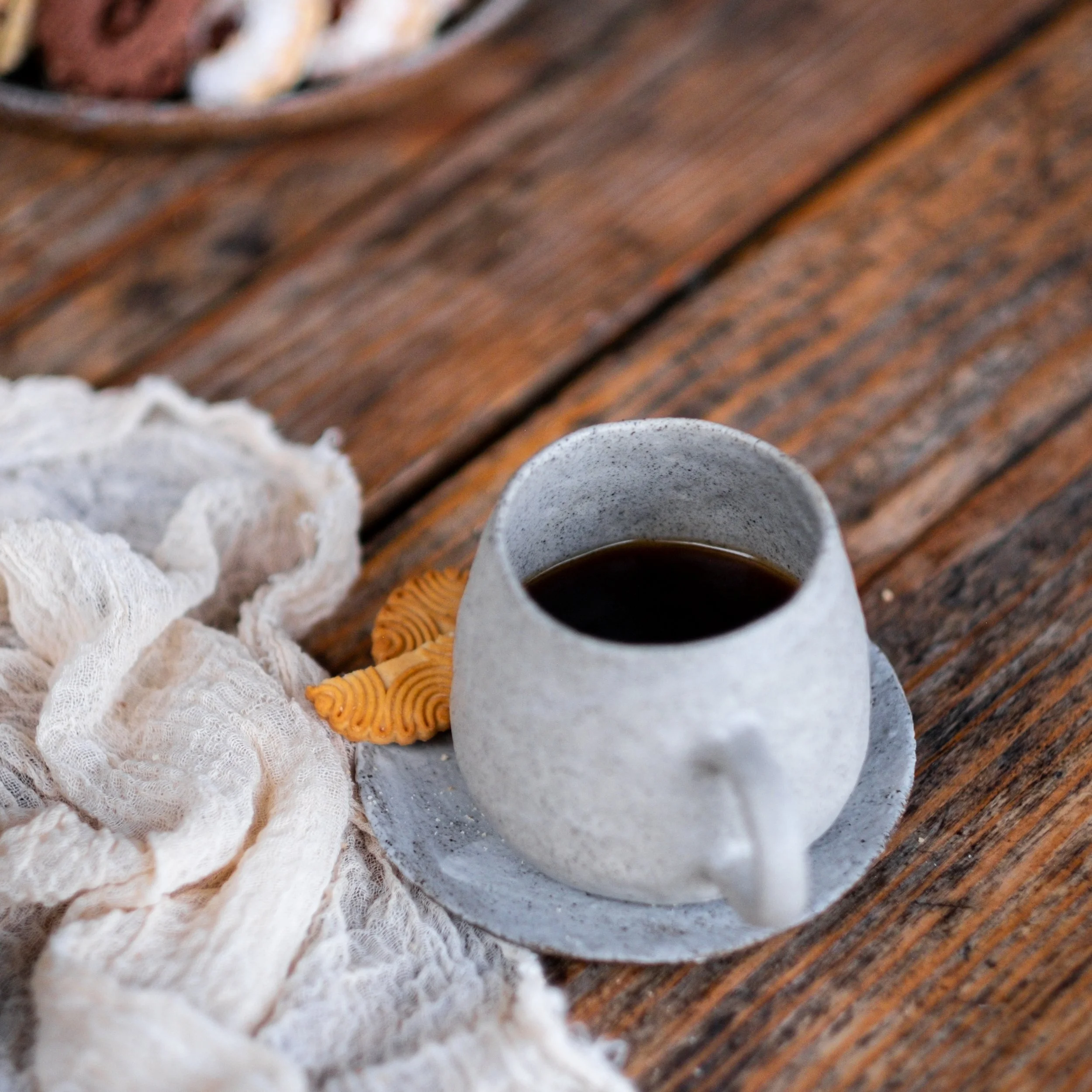 A white ceramic mug filled with black coffee sits on a matching saucer on a rustic wooden table. Next to the mug, there are two orange cookies with a spiral pattern, and a crumpled white cloth drapes nearby.