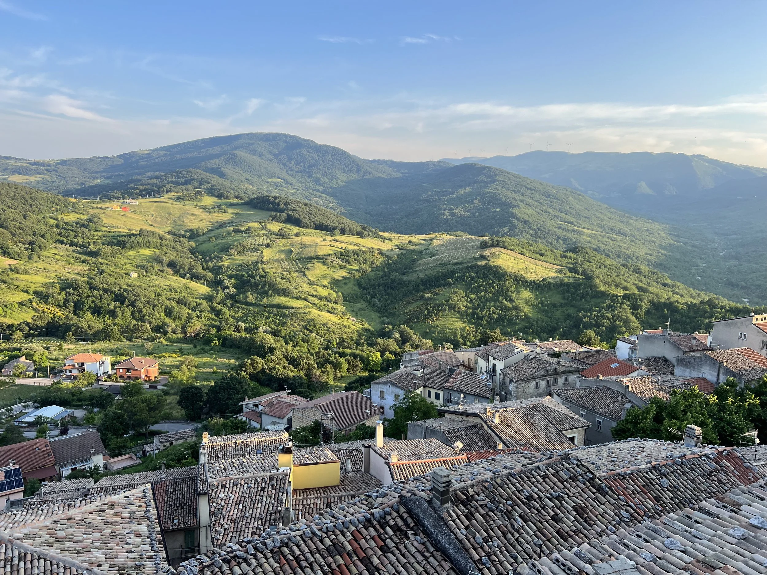 Scenic view of a small village with tiled rooftops in the foreground, green hills and mountains in the background, under a partly cloudy sky.