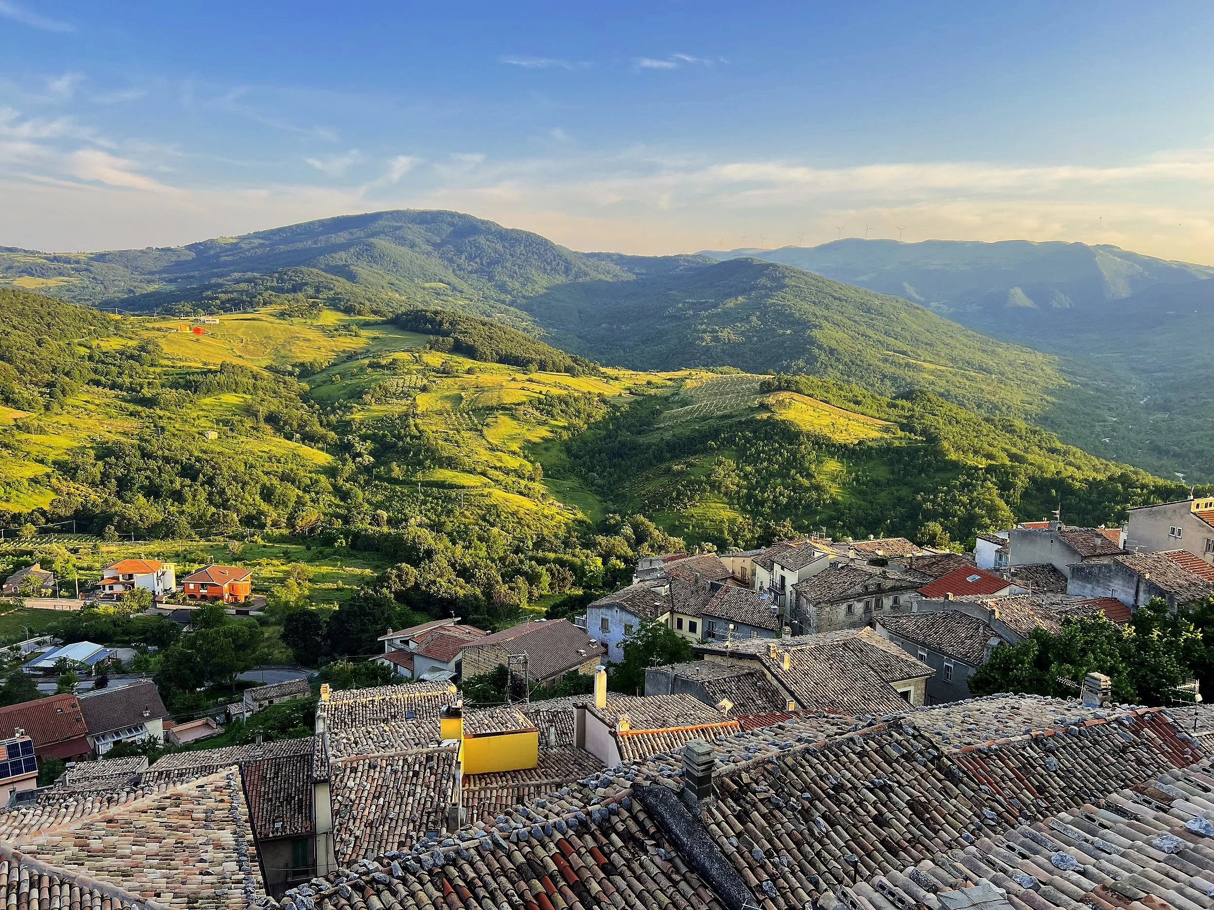 A scenic view of a small town with tiled rooftops in the foreground, surrounded by lush green hills and mountains under a partly cloudy sky.