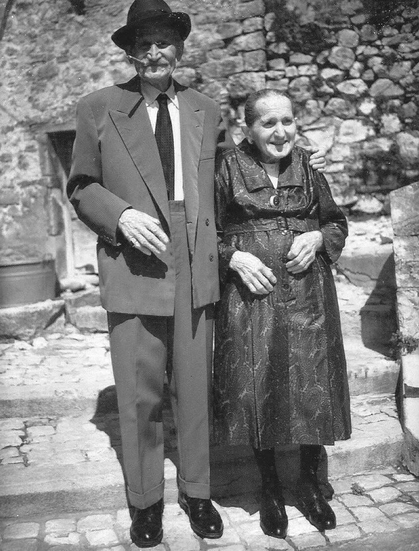 A black-and-white photo of an elderly man and woman standing outdoors on cobblestone steps, smiling and posing for the camera. The man is wearing a suit, tie, glasses, and a hat, and the woman is wearing a patterned dress with a belt. The man has his arm around the woman's shoulder.