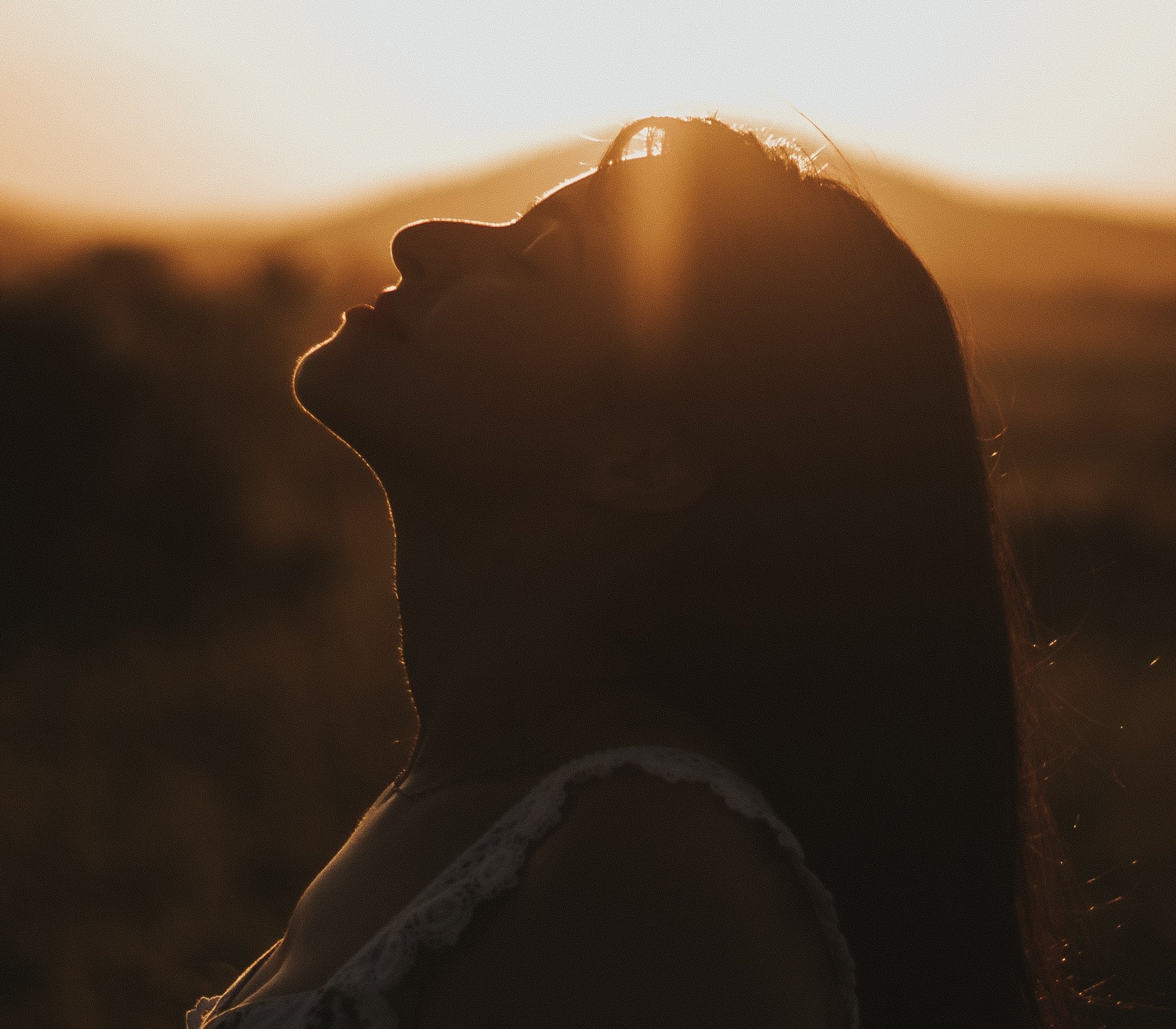 Silhouette of a person sitting outdoors at sunset, with a view of the horizon and warm sunlight casting a glow around the person's profile.