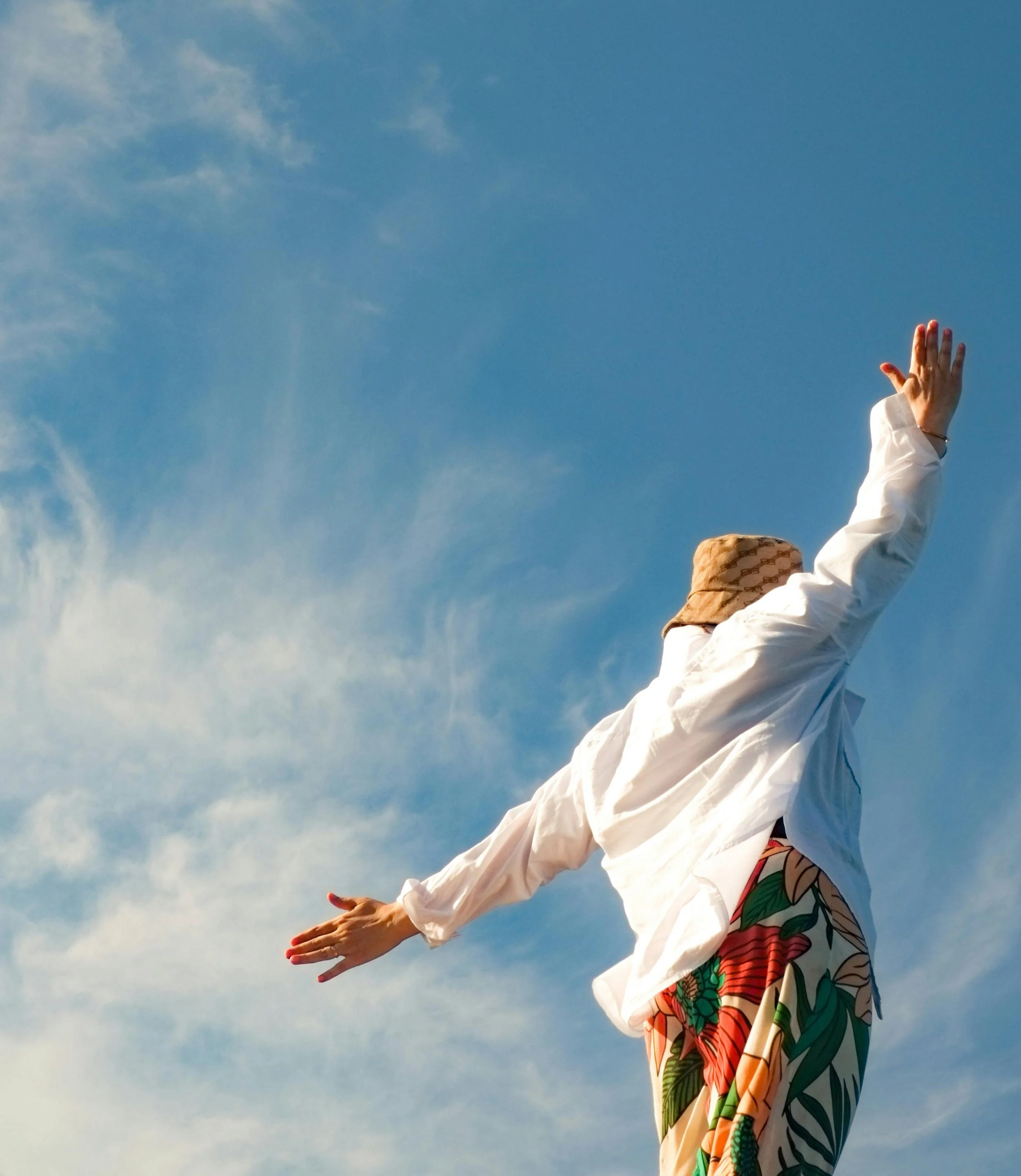 Person wearing a white shirt and patterned skirt reaching out towards the sky with arms extended, against a background of blue sky with scattered clouds.