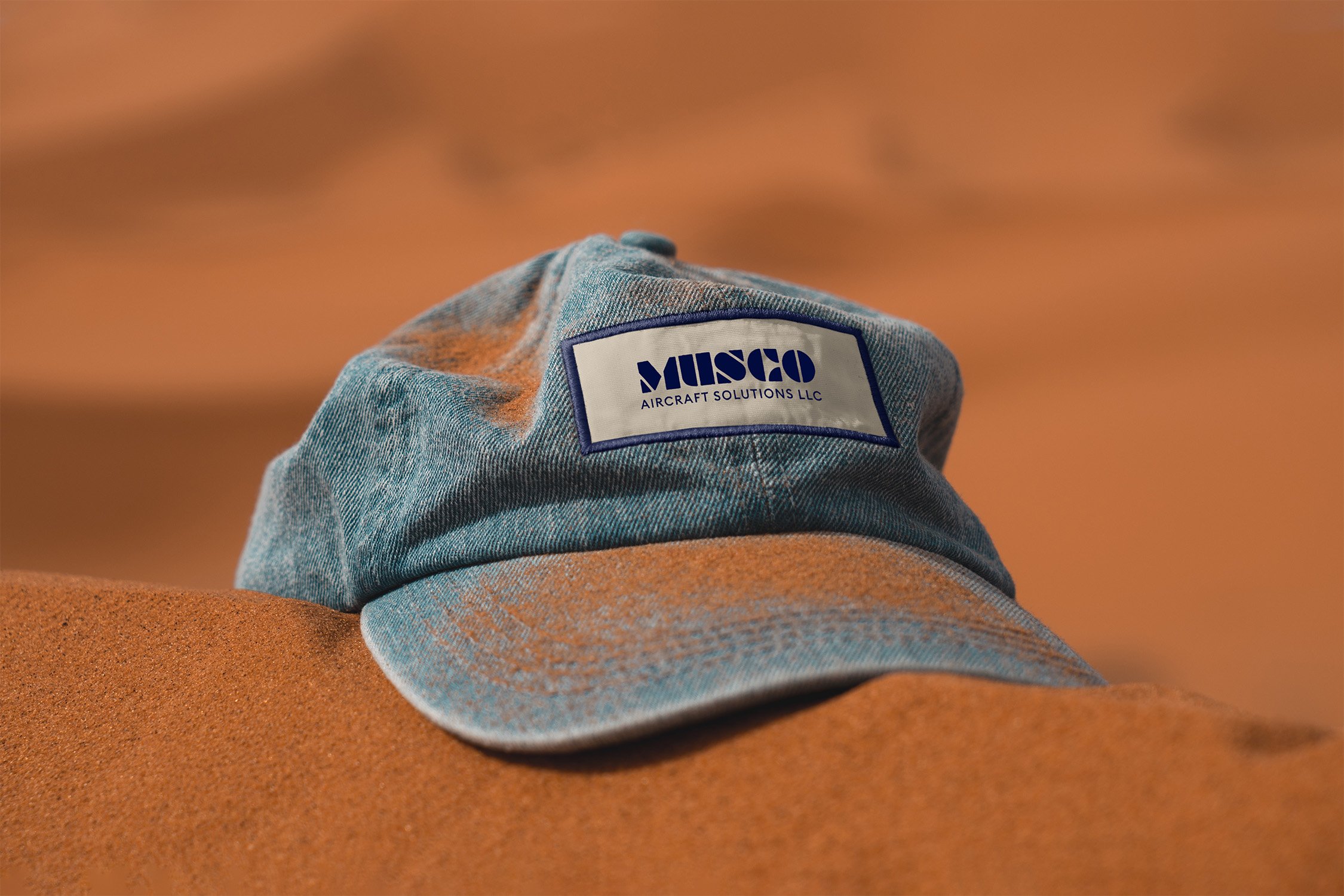 Graphic design mockup of a hat in the sand for Musco Aircraft Solutions — modern, geometric wordmark logo with a clean brandmark with a clean sans serif typography, designed by Will Lefkovich.