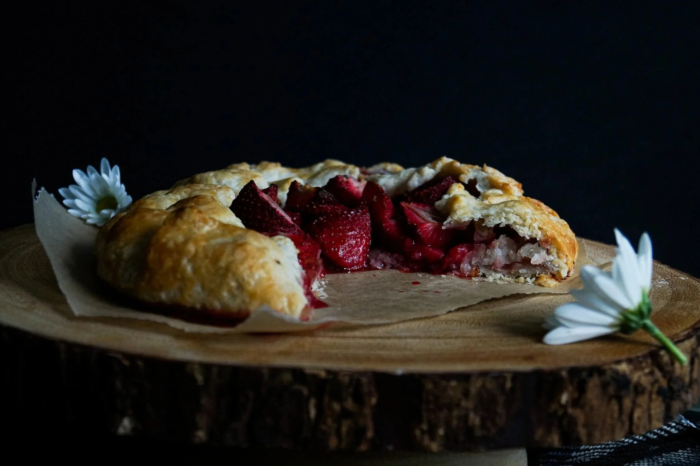 A partially eaten strawberry galette on a wooden serving board with white flowers on the side, against a dark background.