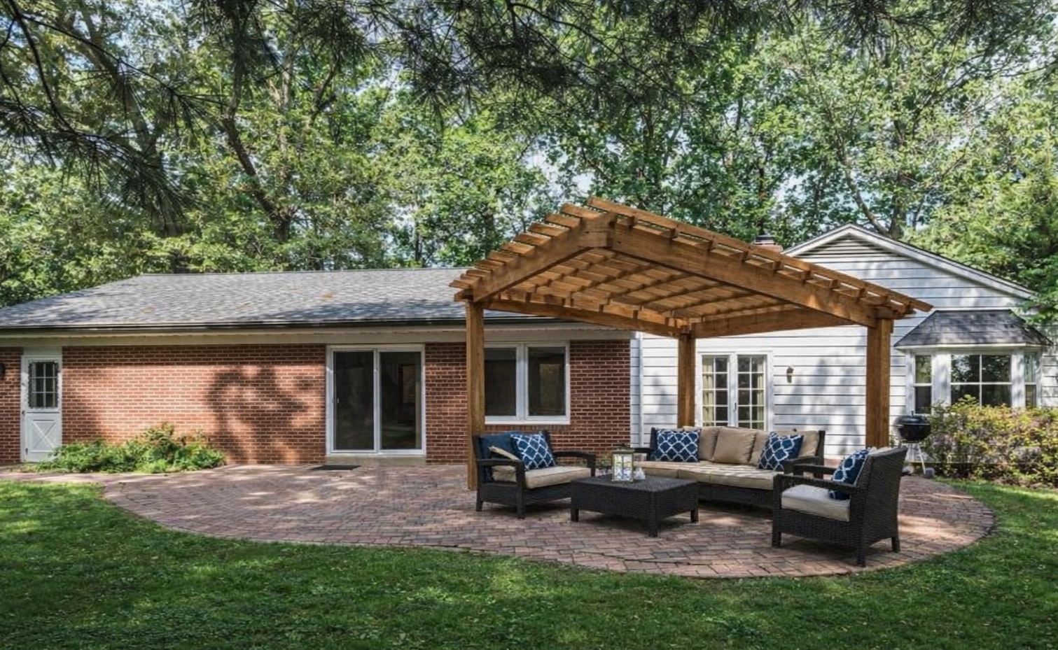 Backyard patio with brick flooring, outdoor seating under a wooden pergola, and a suburban house with white siding and a brick wall.
