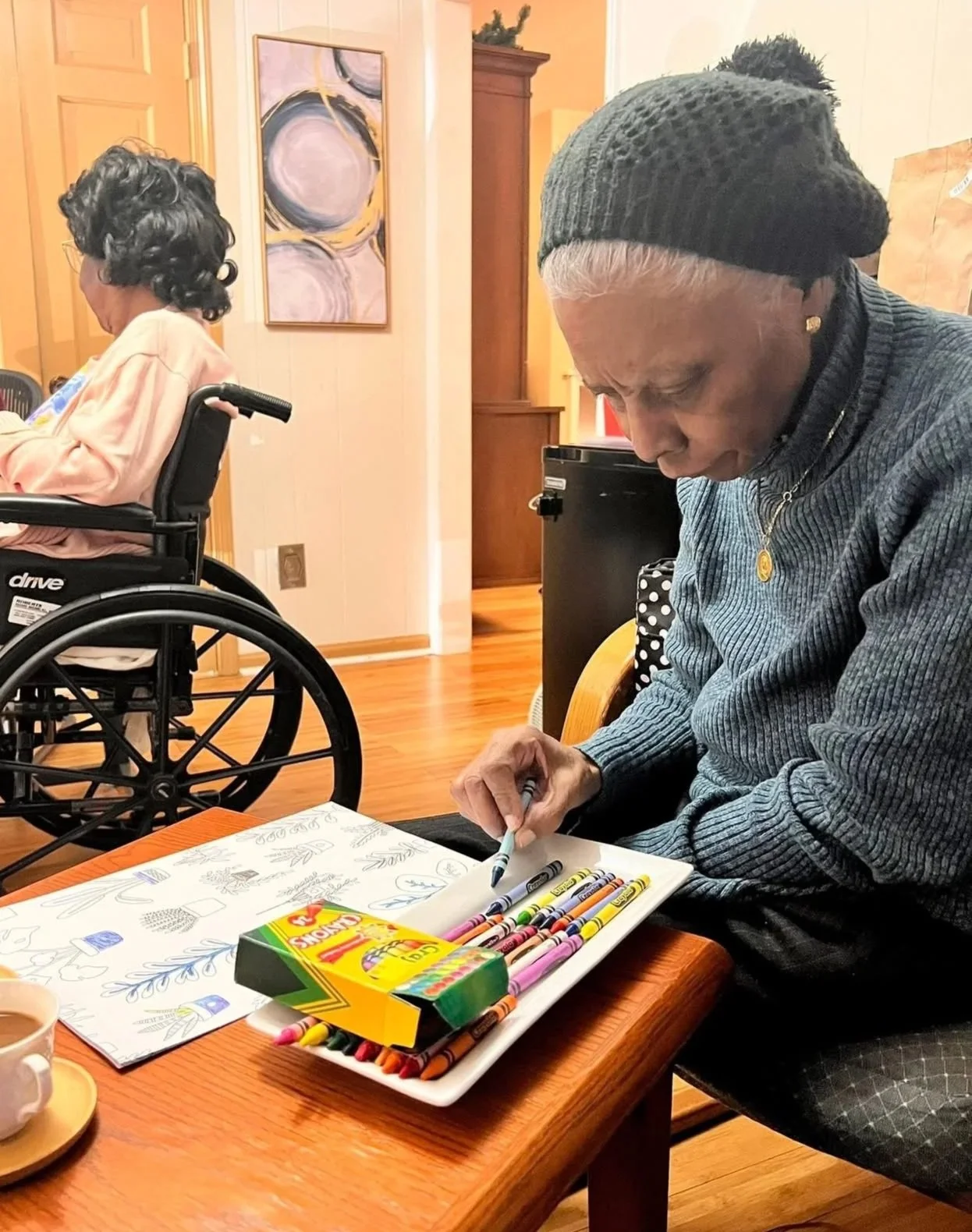 An elderly woman is seated at a table with coloring supplies, including crayons and markers, in front of her. She is wearing a dark knit hat and a dark sweater. In the background, a woman in a wheelchair with black curly hair and a light pink sweater