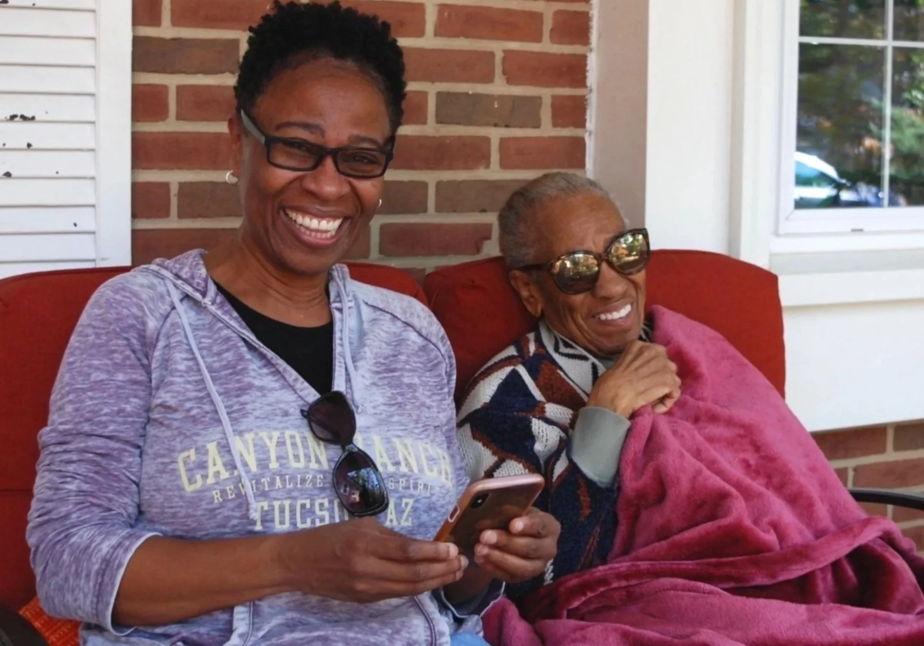 Two women sitting on a porch swing, smiling and enjoying themselves. The woman on the left is holding a smartphone and wearing a gray hoodie and sunglasses, and the woman on the right is an elderly woman wearing sunglasses and a multicolored sweater,