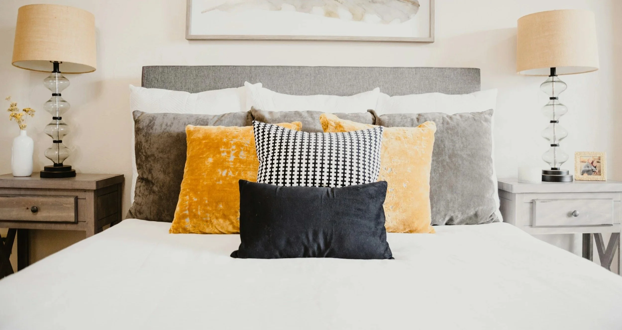 A neatly made bed with a gray headboard, decorated with multiple pillows in black, gray, yellow, and white with black pattern, flanked by two nightstands with lamps, and framed artwork hanging above.