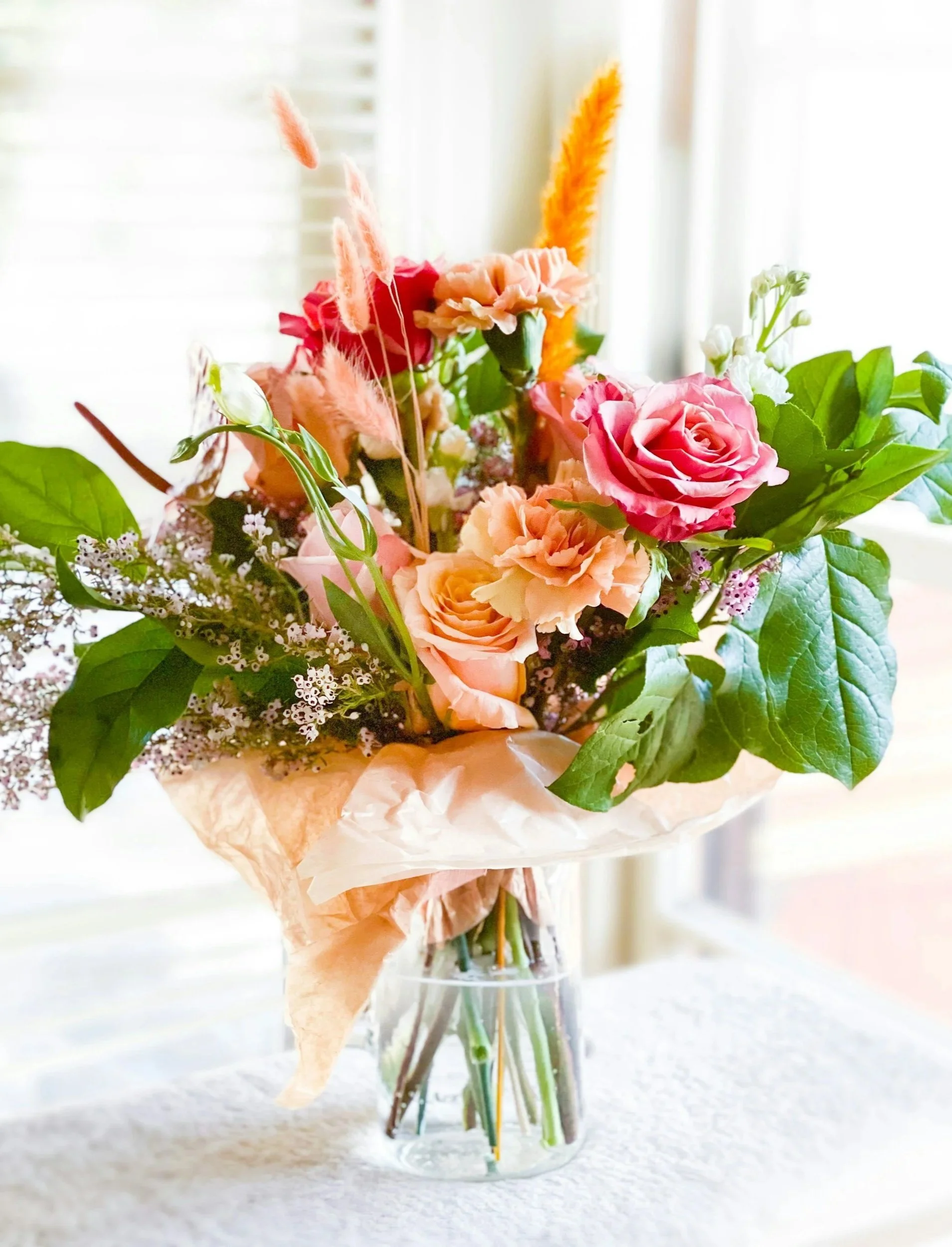 Vase with pink and peach roses, greenery, and dried flowers on a table near a window.