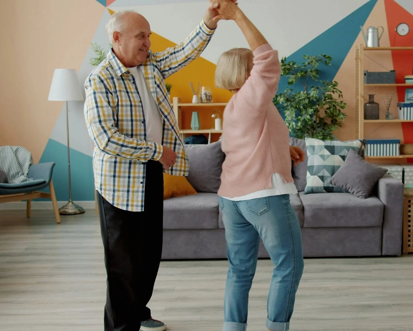 An elderly man and a middle-aged woman dancing together in a living room. The man is holding the woman's hand as she spins, smiling and enjoying the moment. The living room has a gray sofa with pillows, a bookshelf, and a colorful, geometric wall pat