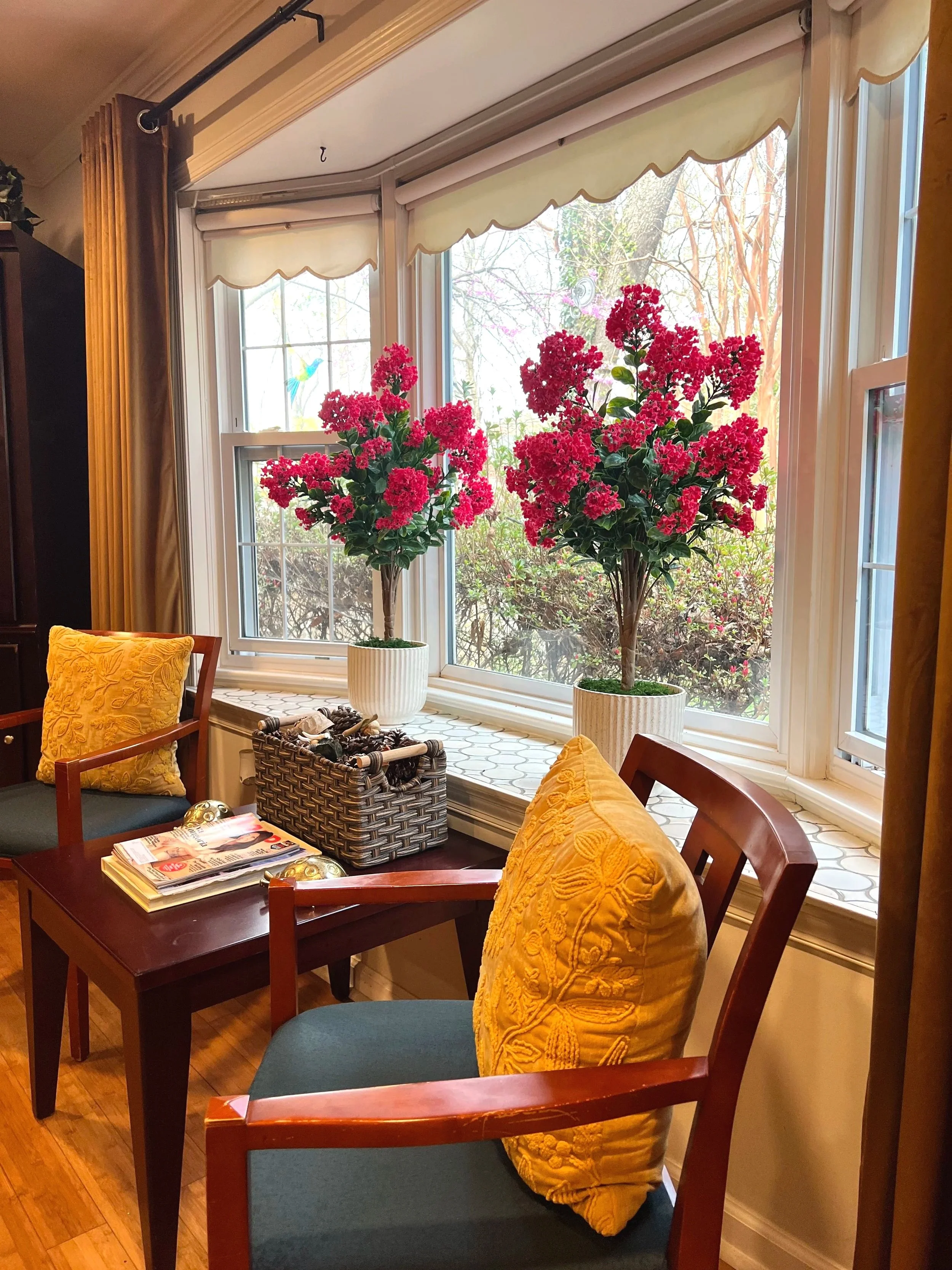 A cozy sitting area with two wooden chairs with upholstered cushions, a small wooden table between them, and a large bay window with cream-colored curtains. The window has two hanging flower baskets, a potted plant, and a basket of small pumpkins on the windowsill. Outside, there is lush greenery and trees.