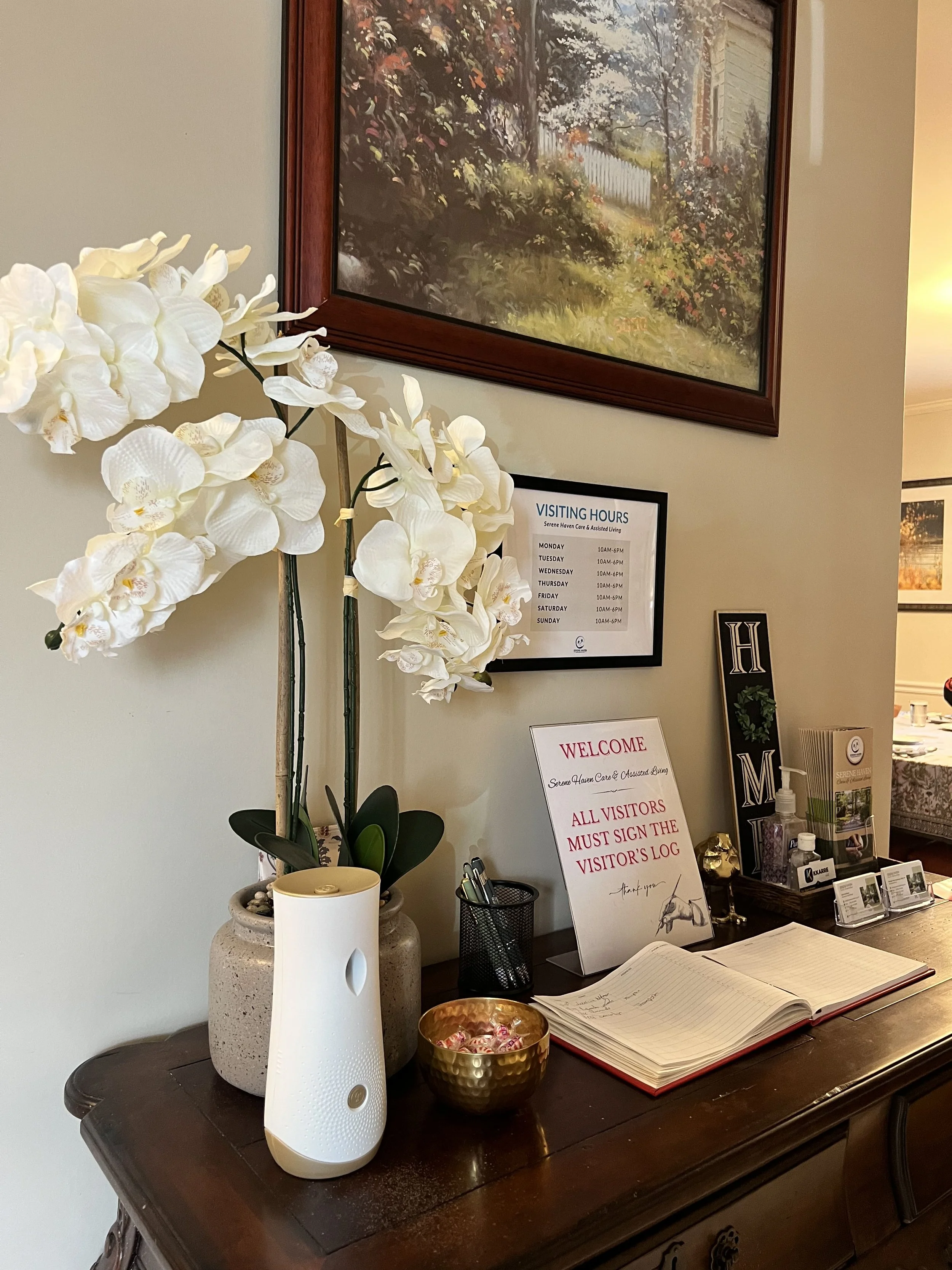 Dining room with a dark wooden table set with orange plates, surrounded by six orange chairs, and a large potted plant centerpiece. There are windows and artwork on the walls, and a doorway leading to a living area with red armchairs.