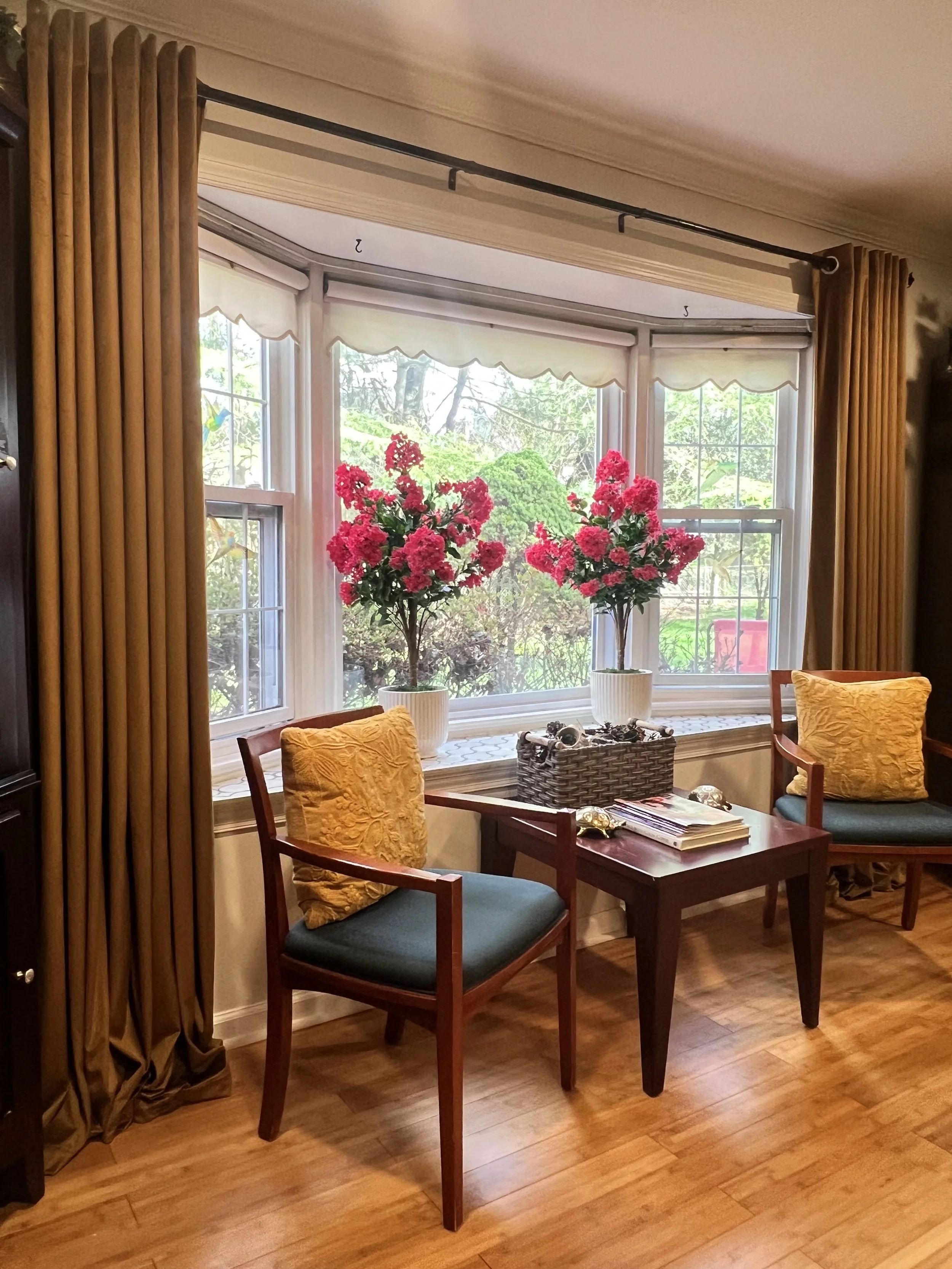 A cozy living room corner with two dark blue armchairs facing each other, a coffee table in between, decorated with a bouquet of white flowers, a small pumpkin, and books. Behind the chairs is a large window revealing green foliage outside, decorated with orange autumn leaf stickers.