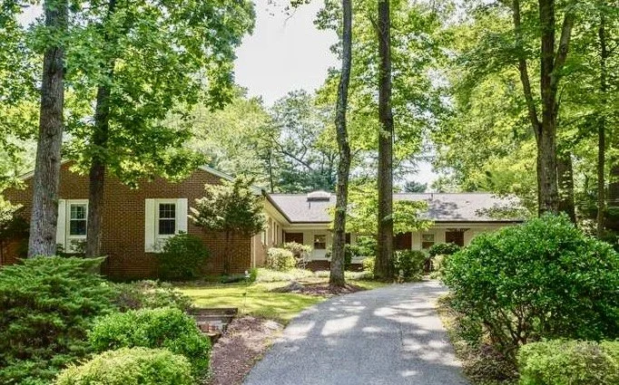 A brick house surrounded by lush green trees and shrubs with a concrete driveway leading to the entrance.