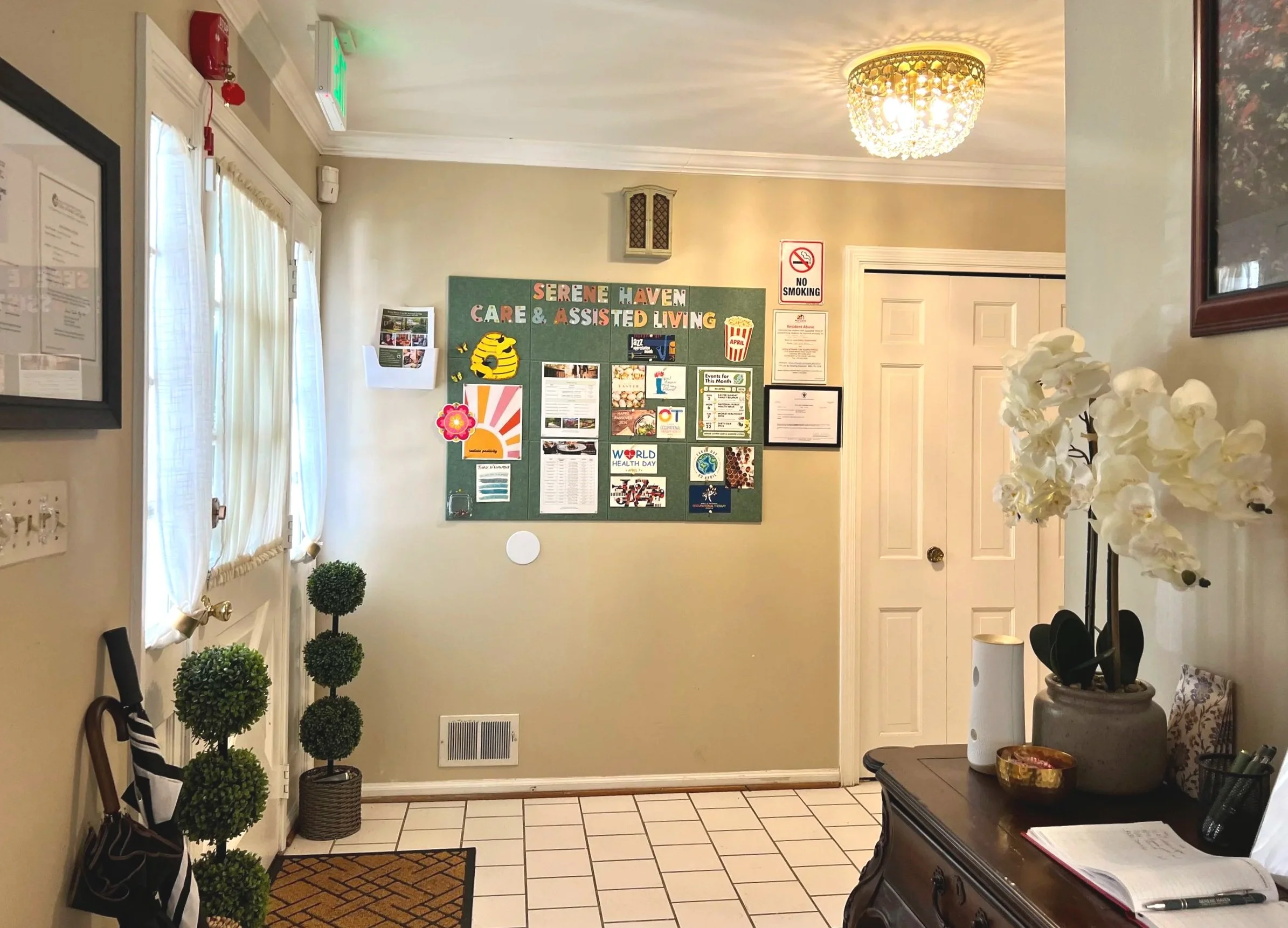 Residential hallway with a white door decorated with a flower wreath, framed pictures on the wall, a small potted topiary plant, and a wooden console table with decorative items and papers, illuminated by wall-mounted lights.