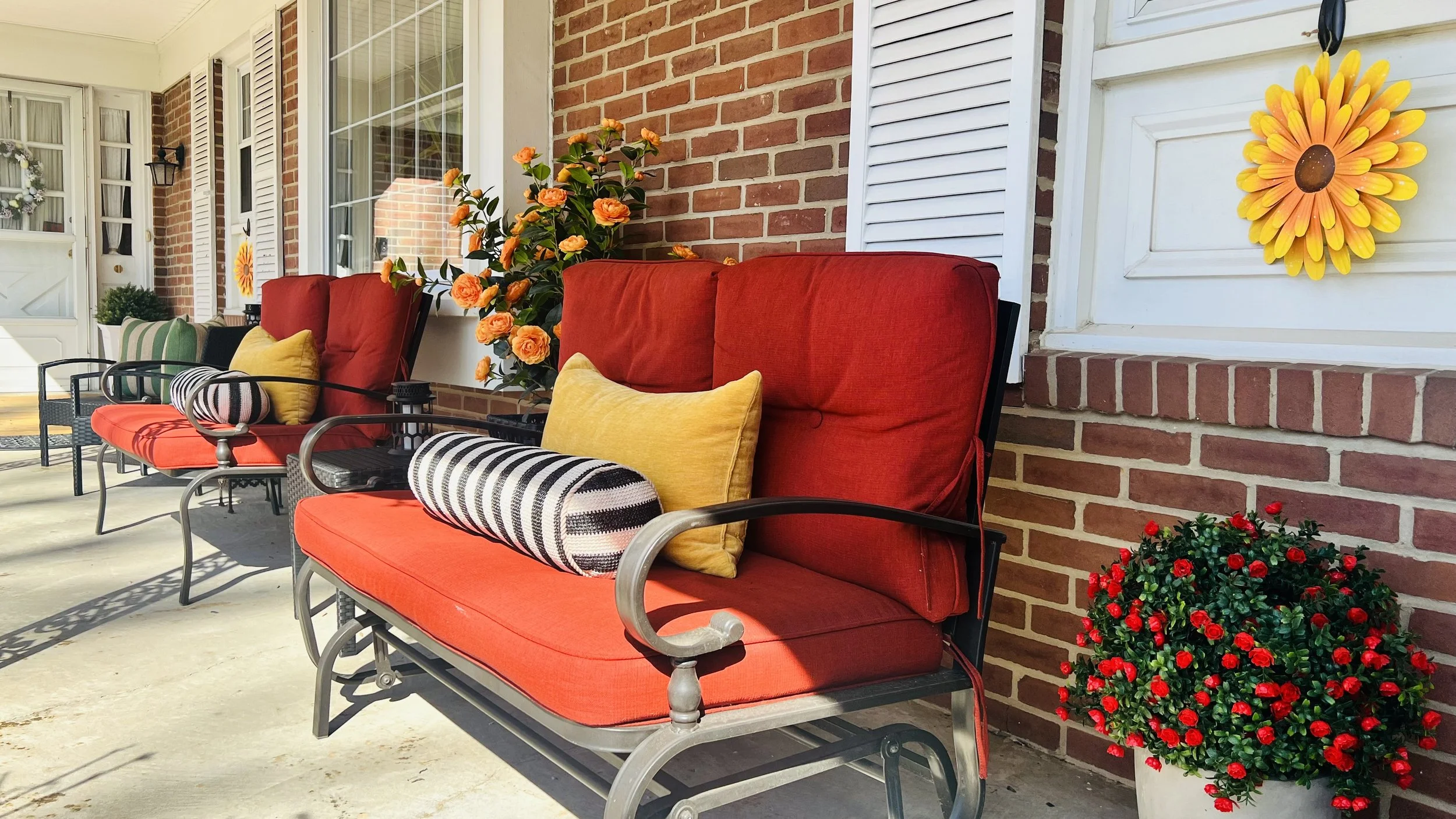Red cushioned outdoor seating with throw pillows, potted plants, and decorative lanterns on a porch with brick and white siding.