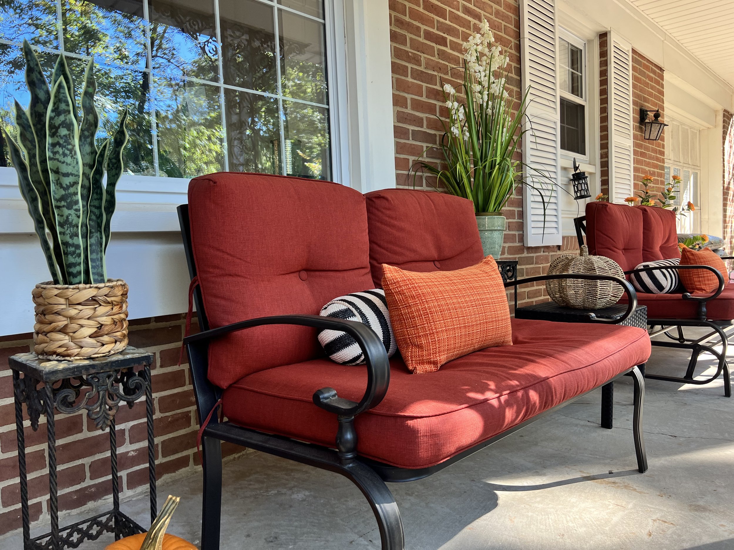 Red cushioned outdoor seating with throw pillows, potted plants, and decorative lanterns on a porch with brick and white siding.