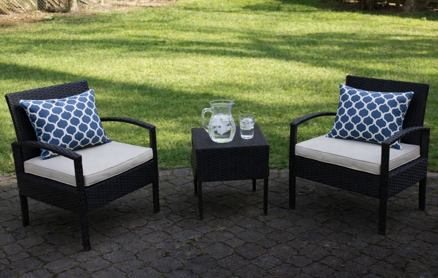 Two black wicker outdoor chairs with white cushions and blue patterned pillows facing each other, separated by a small black wicker table with a glass pitcher and glass of water, set on a stone patio with a green lawn in the background.