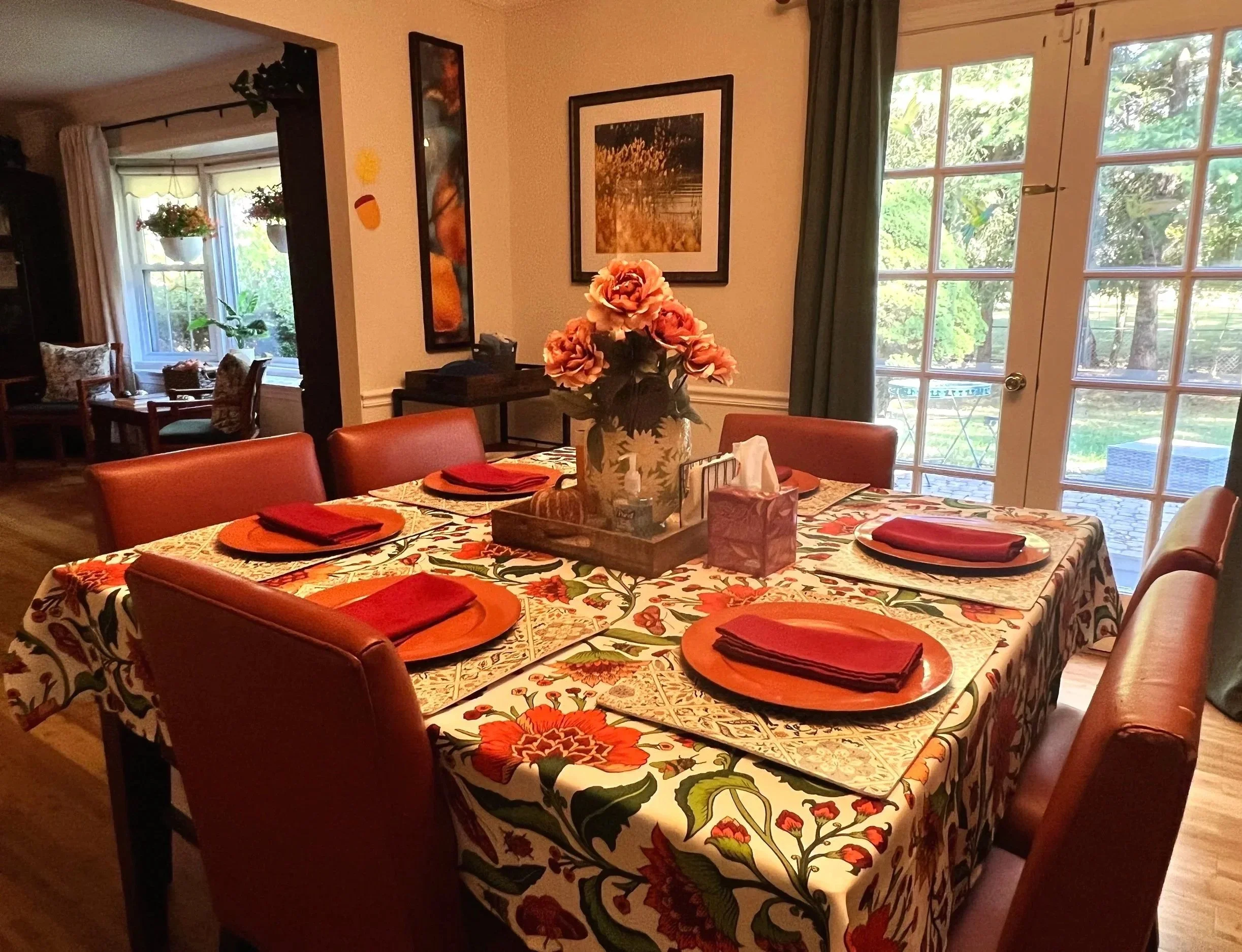 Dining room with six orange plates on a floral tablecloth, a green potted plant in the center, windows with curtains, framed artwork on the walls, and a door to the outside.
