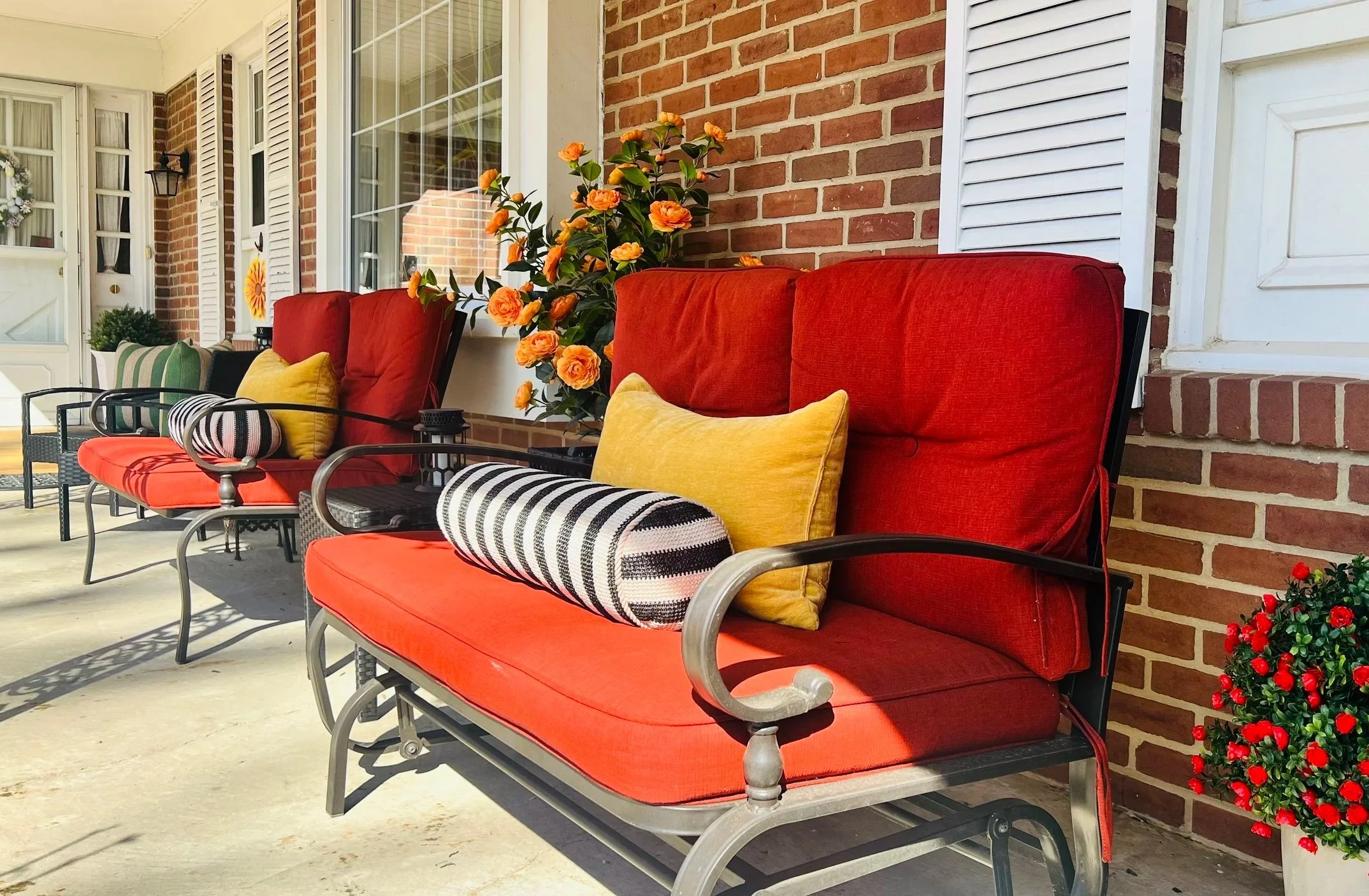 Red cushioned patio chairs with orange pillows on a porch decorated for fall, with pumpkins, a small pumpkin-shaped decorative piece, a woven pumpkin ornament, and a plant in a black pot, next to a brick house with a wreath on the door.